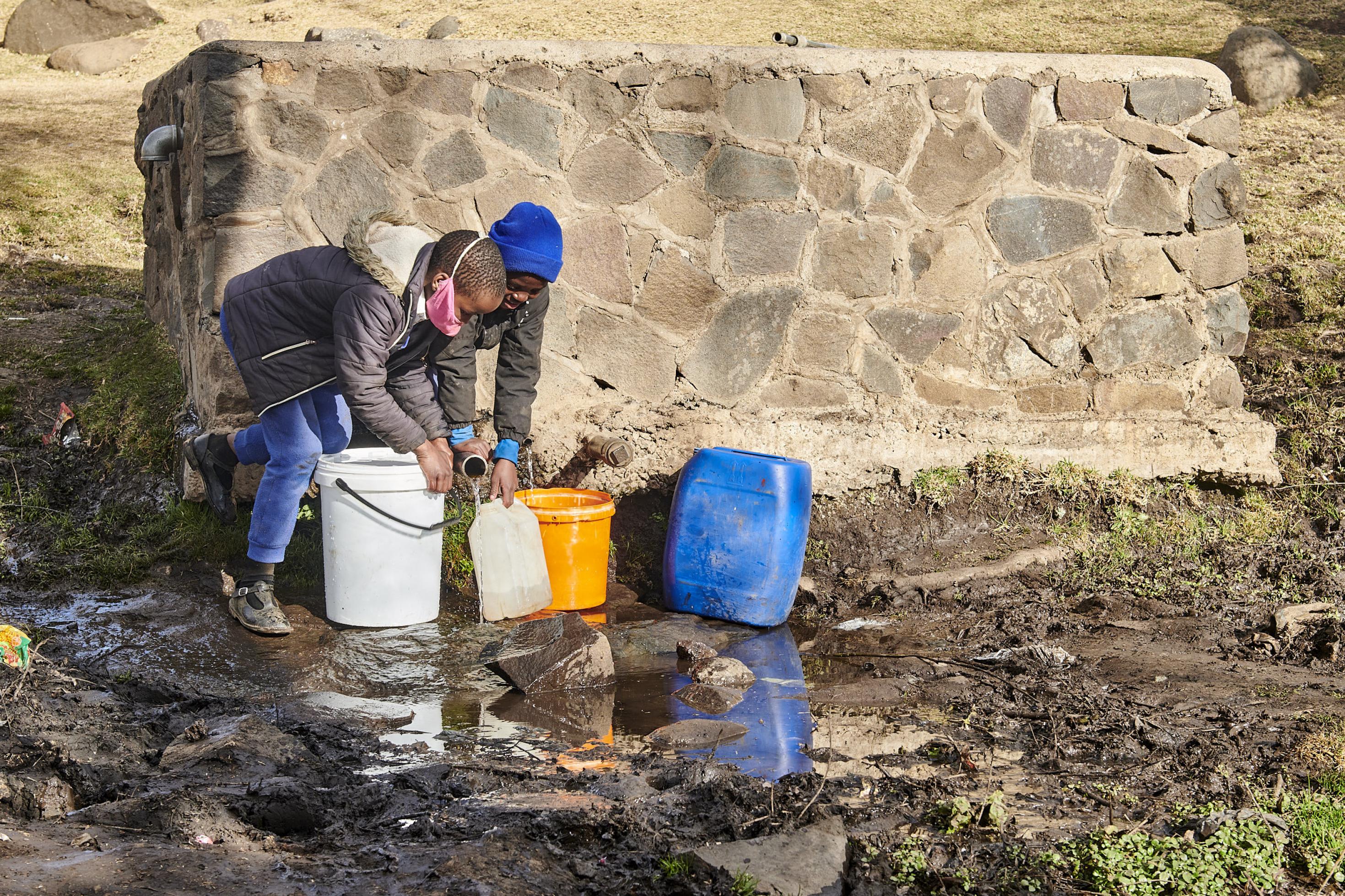 Zwei Mädchen stehen mit mehreren Wasserkanistern an der Wasserquelle – einem Steinquader aus dem zwei Rohre ragen