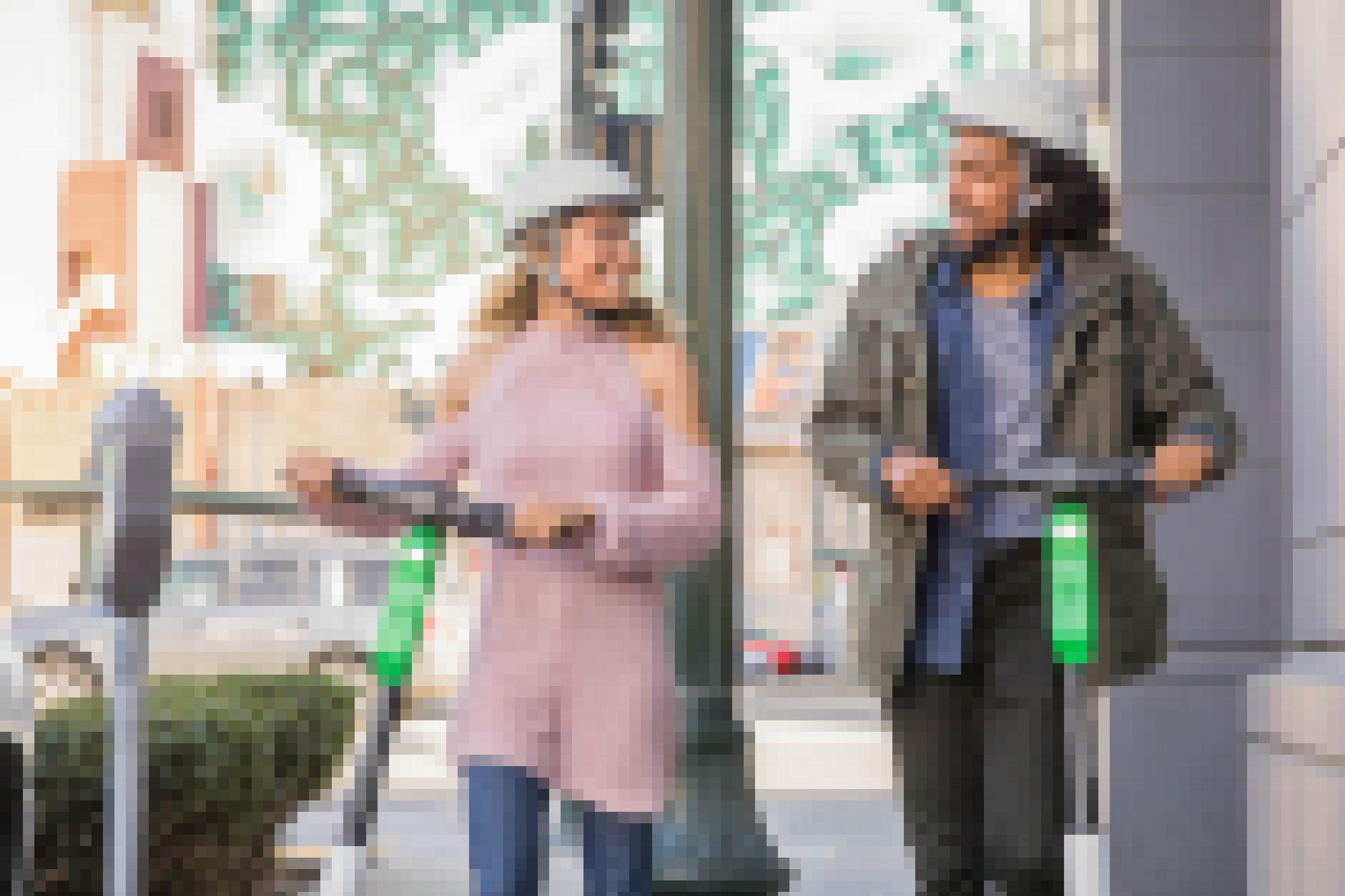 Eine Frau und ein Mann mittleren Alters mit jeweils einem E-Scooter sind in der Stadt unterwegs und lächeln sich an. Sie tragen beide einen Helm.