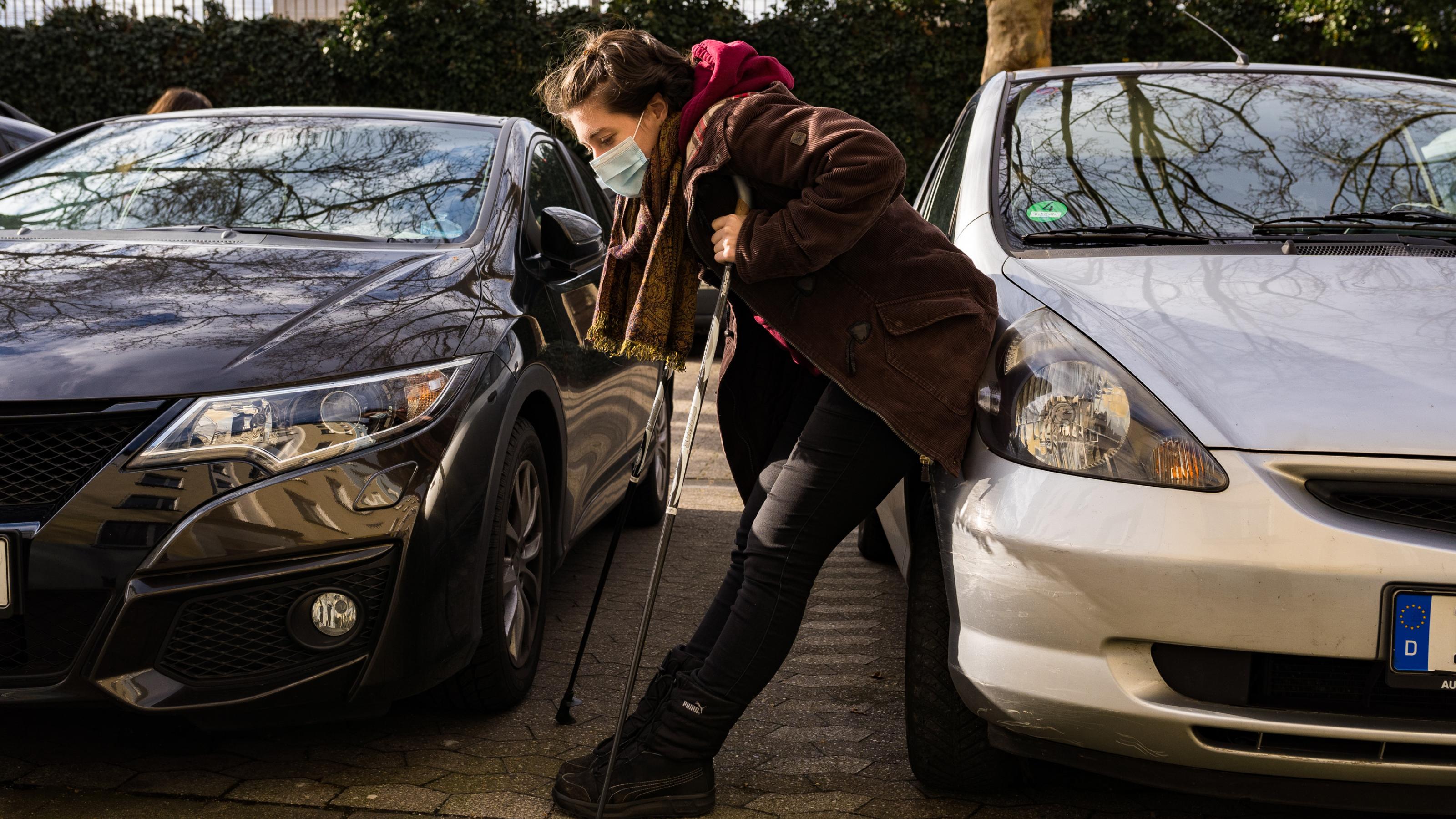 Luisa Meißner steht gebückt auf Krücken zwischen zwei Autos, und ruht sich aus. Sie hat eine Atemschutzmaske auf.