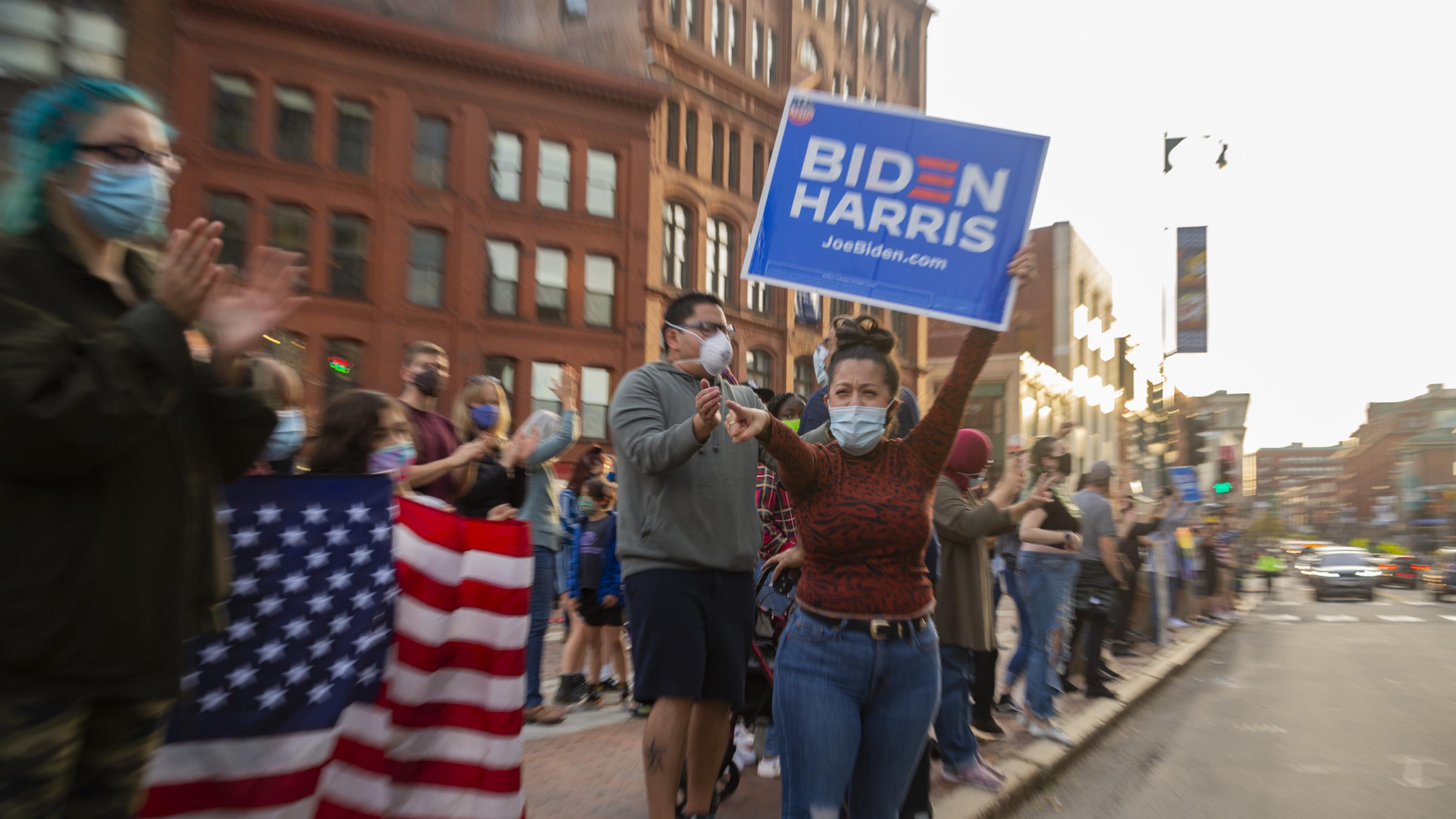 Eine Gruppe von maskentragenden Menschen steht in einer Innenstadt am Straßenrand. Eine Frau hält ein Biden-Plakat in die Höhe.