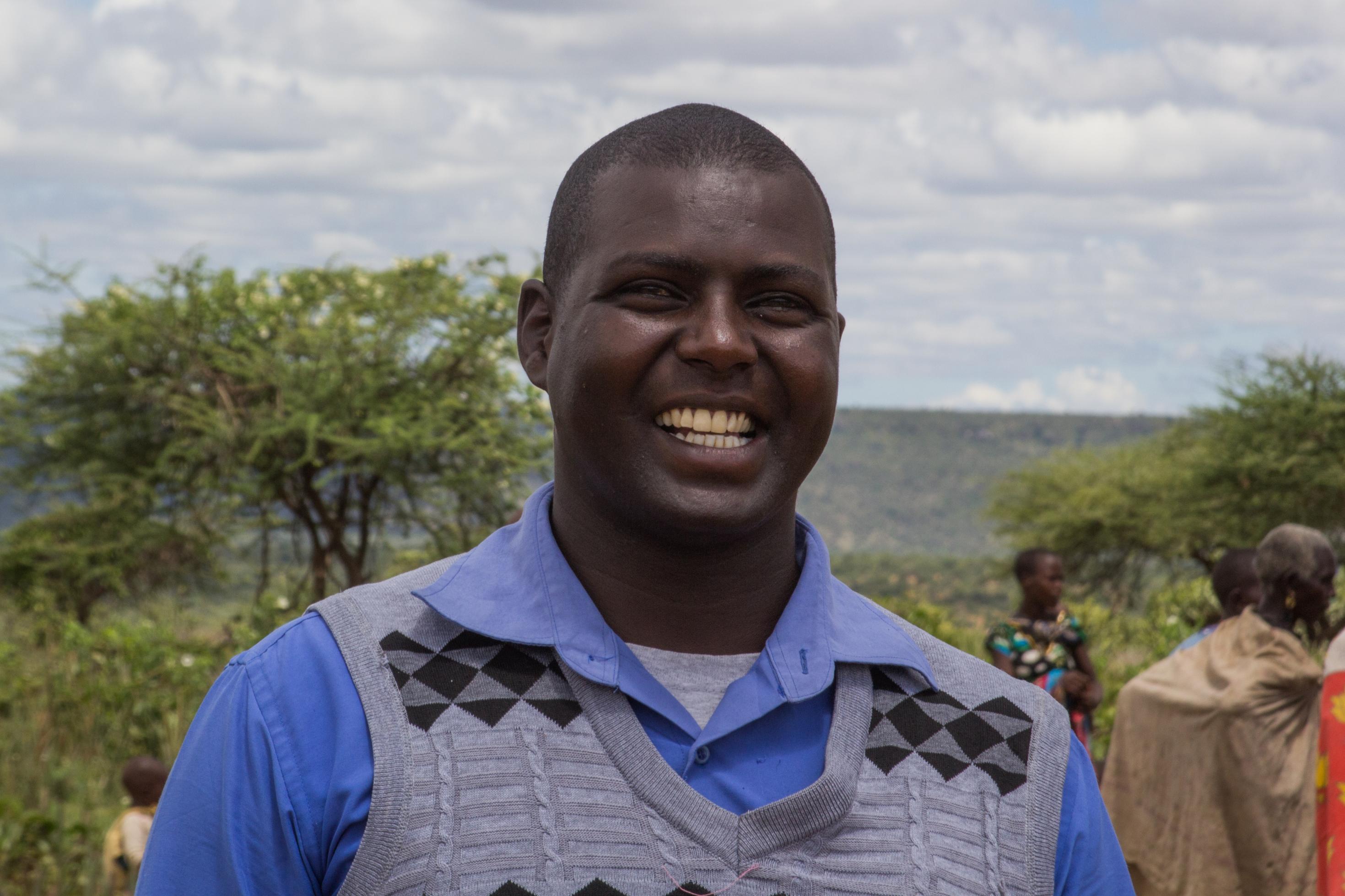 Massai-Chief Stephen Lesengoi am Rande der Verteilung der Lebensmittel. Auf dem Bild ist ein Mann Mitte 30, er trägt einen Pollunder und ein blaues Hemd. Im Hintergrund sind Frauen in traditionellen, bunten Kleider zu sehen.