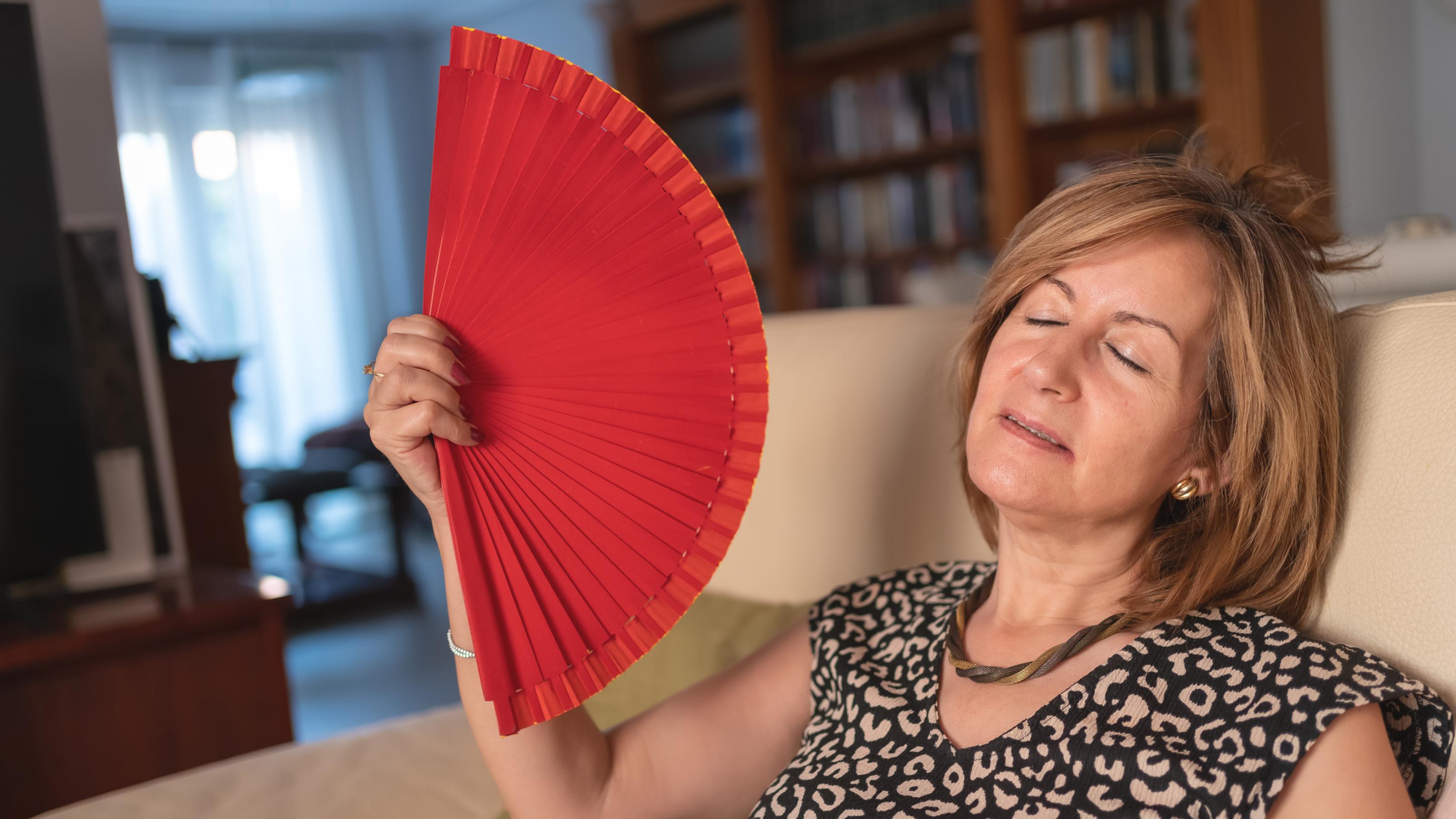 Frau im mittleren Alter, sitzt in ihrem Wohnzimmer auf dem Sofa und wedelt mit einem roten Handfächer vor ihrem Gesicht, um sich abzukühlen
