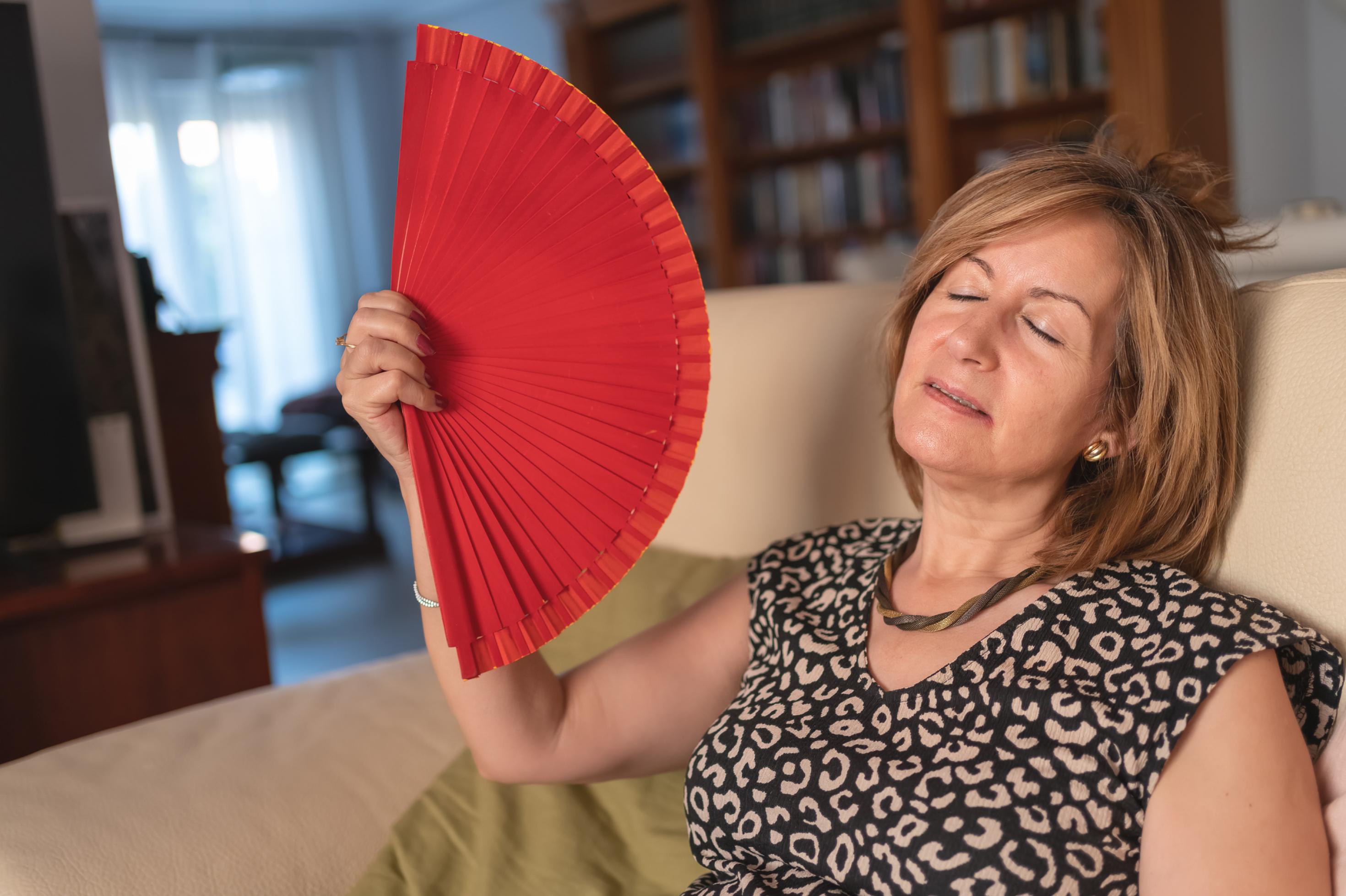 Frau im mittleren Alter, sitzt in ihrem Wohnzimmer auf dem Sofa und wedelt mit einem roten Handfächer vor ihrem Gesicht, um sich abzukühlen