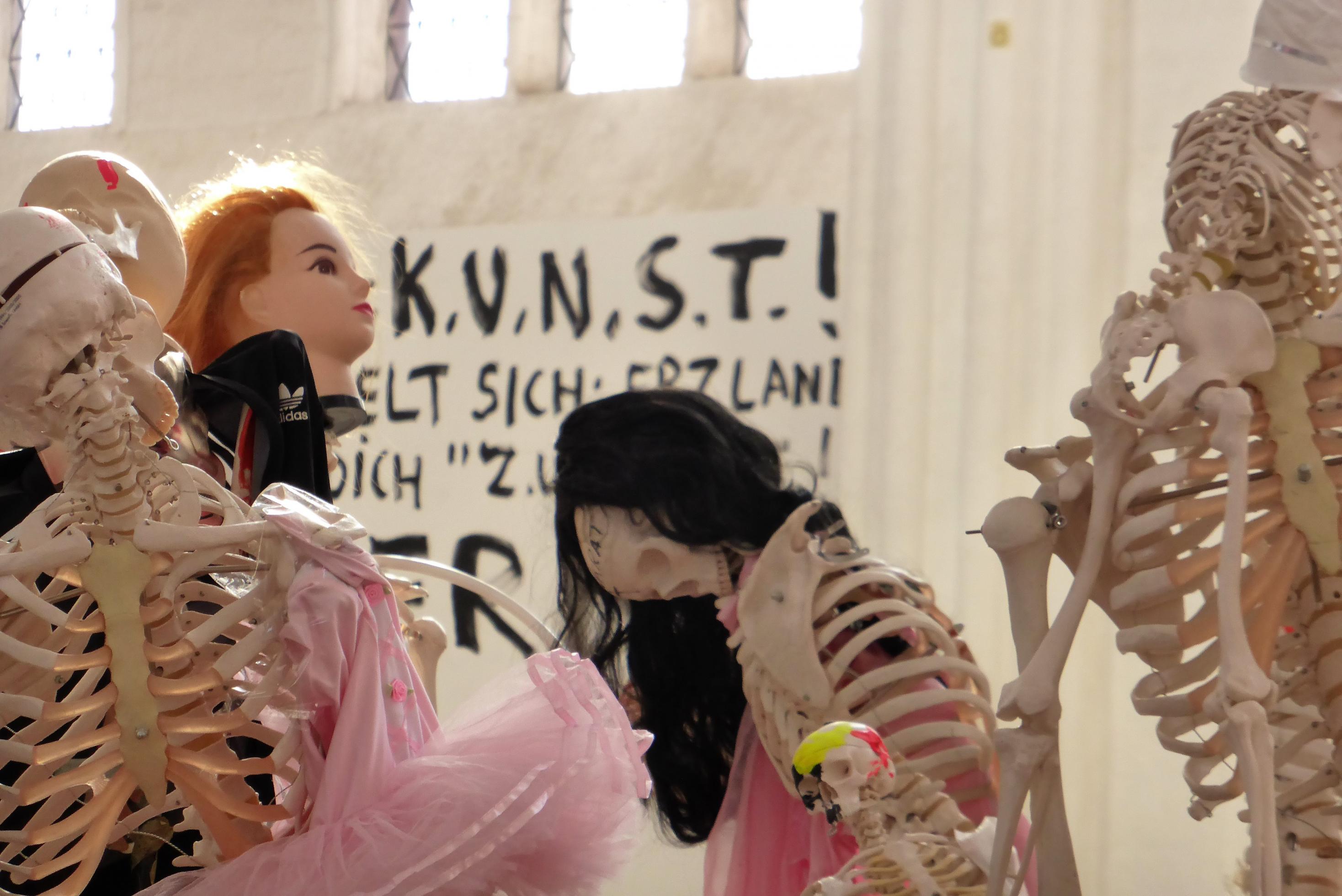 Detail der Meese-Installation: Puppenartig verfremdete Skelette stehen vor einem Wandplakat mit der Aufschrift KUNST!