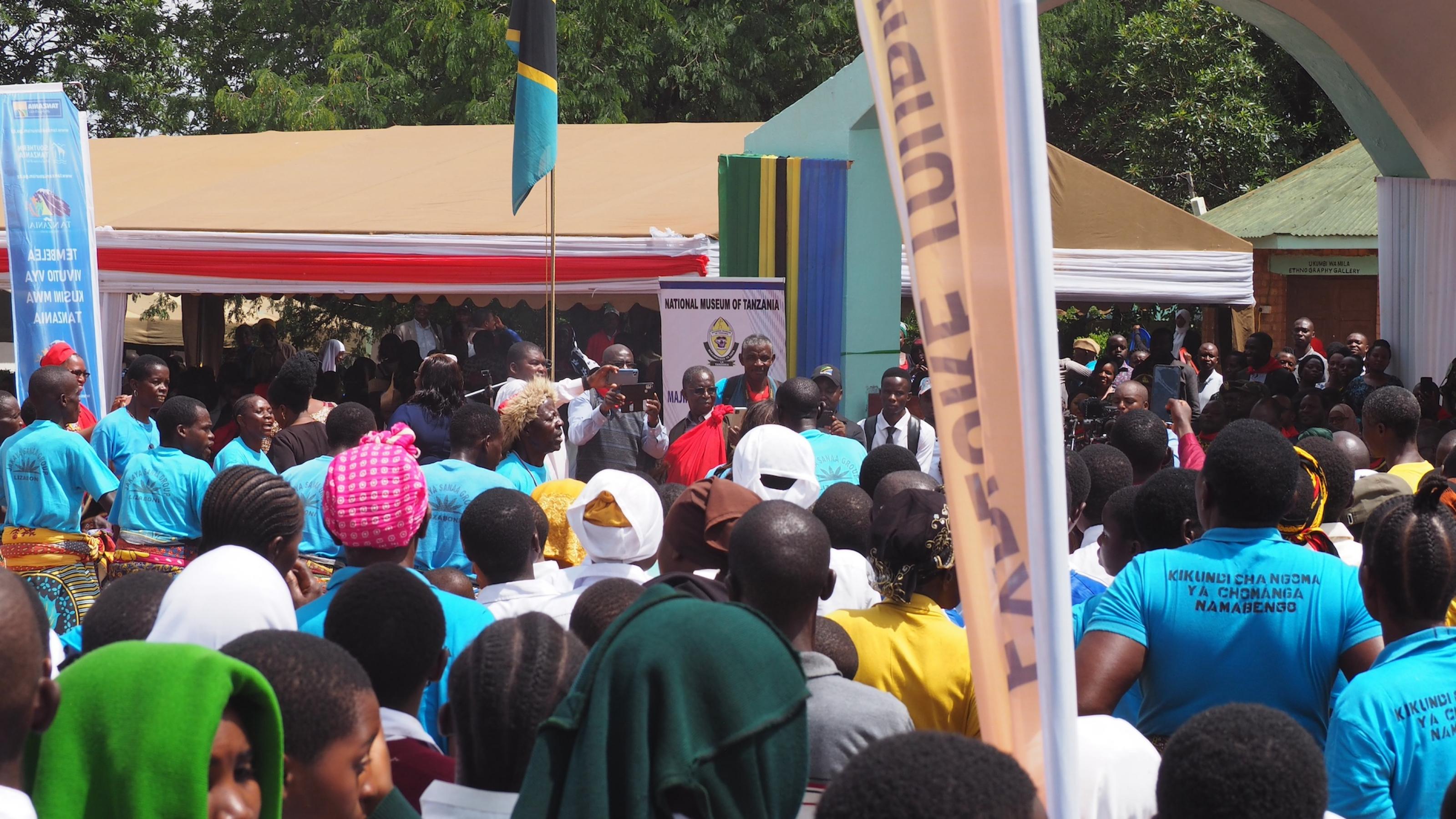 Eine Menschenmenge, zwei tansanische Flaggen, ein Banner mit Aufschrift „National Museum of Tanzania“