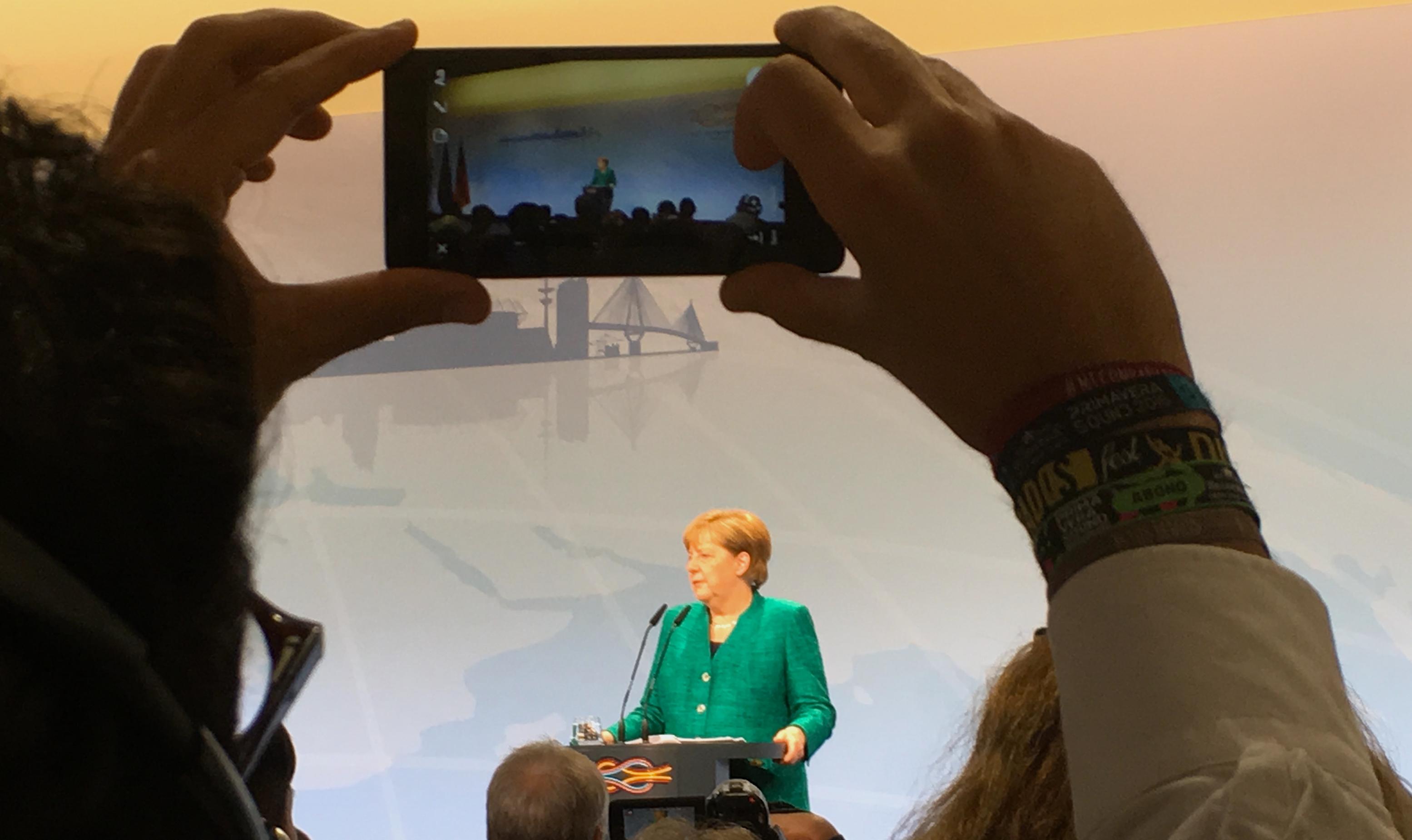 Das Bild zeigt Bundeskanzlerin Angela Merkel auf dem Podium und am Mikrophon bei der Pressekonferenz zur Verkündigung der Ergebnisse des G20-Gipfels in Hamburg. Es ist aus einer hinteren Reihe fotografiert, zwischen den Armen eines anderen Journalisten hindurch, der Merkel gerade mit seinem Smartphone aufnimmt. Man sieht die CDU-Poltikerin noch einmal auf dem Bildschirm des Geräts.