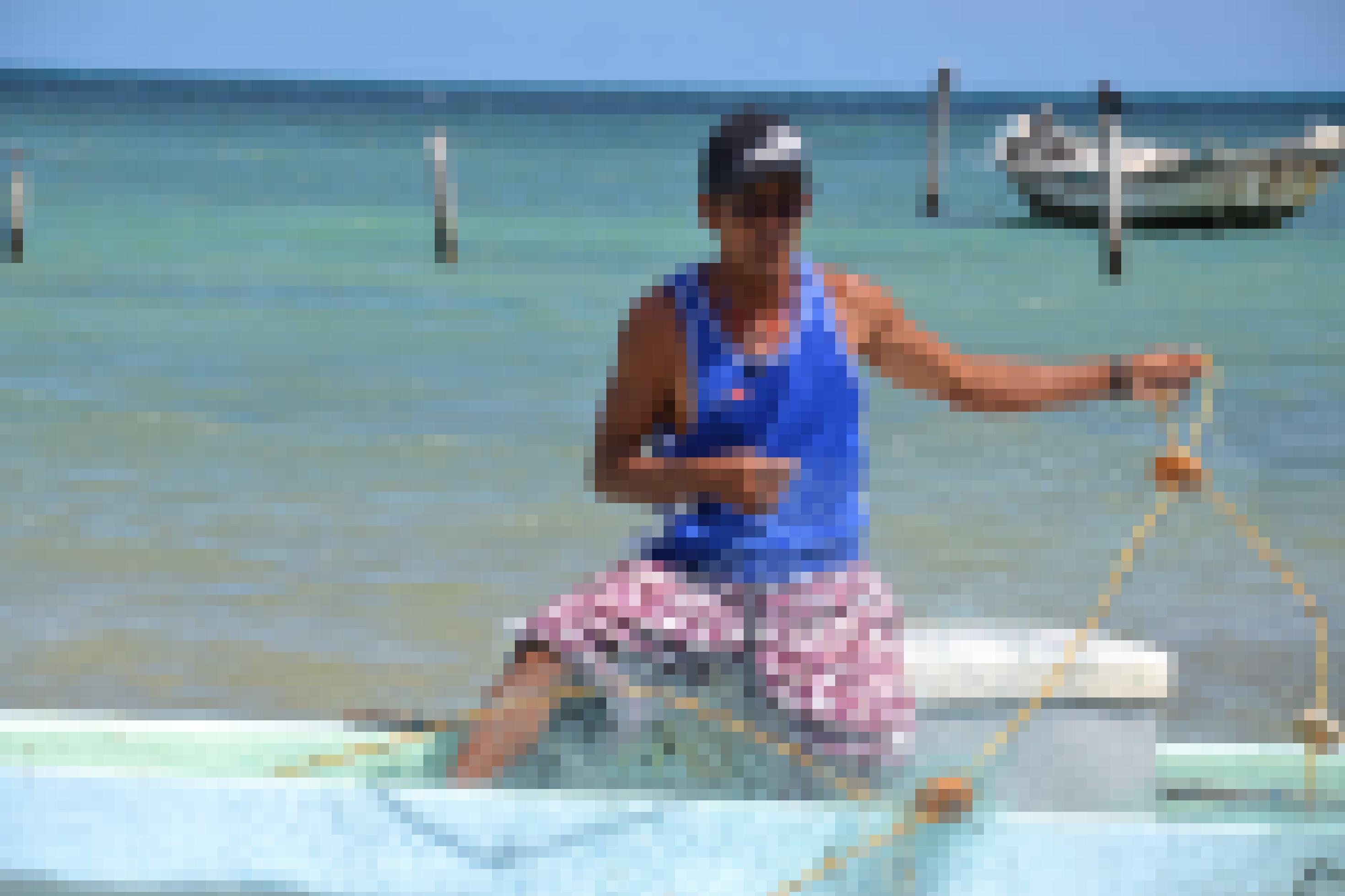 Ein Fischer repariert sein Netz, auf einer Kiste sitzend im Boot, dahinter die türkisblaue Karibik.