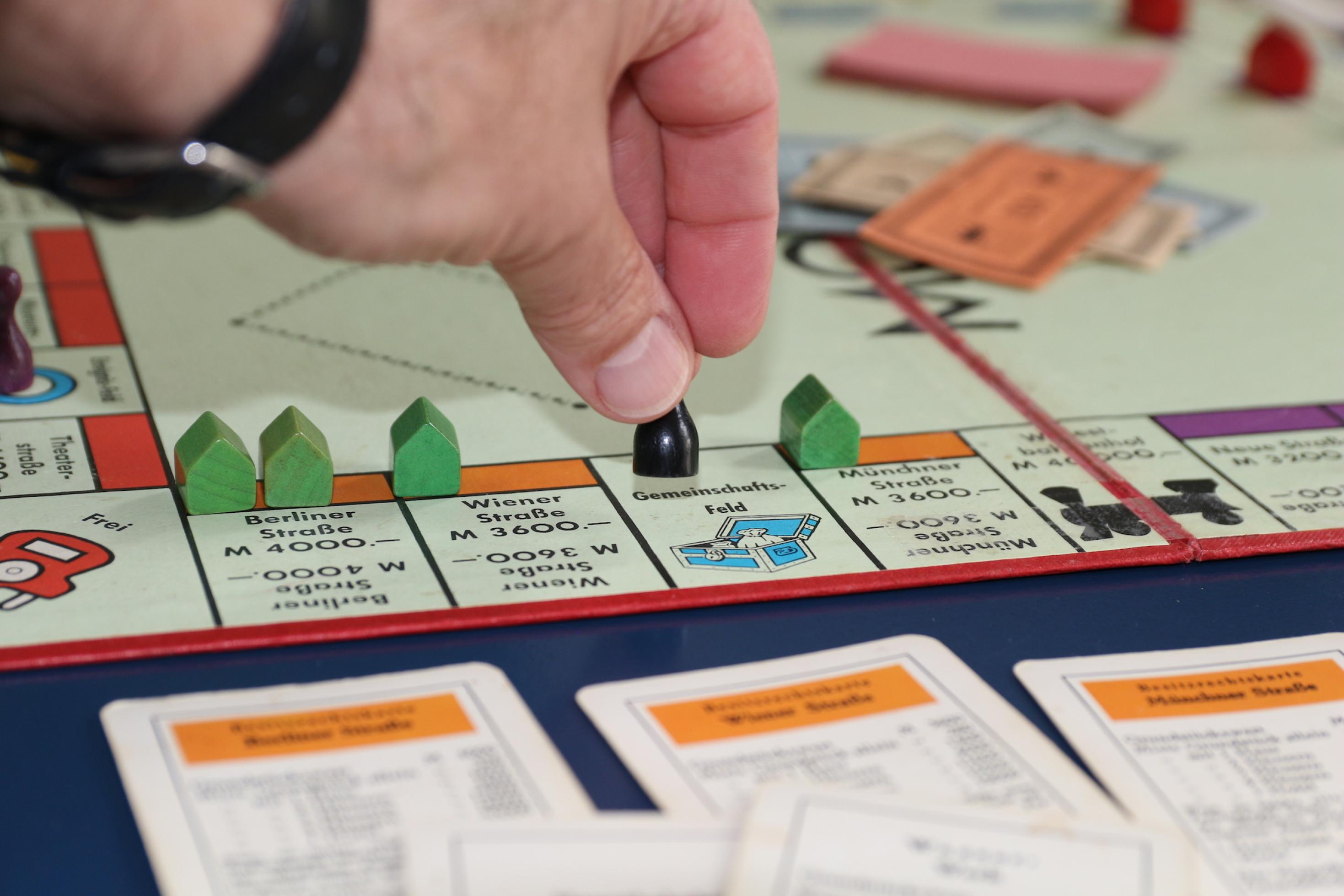 Spielsituation bei Monopoly: Die schwarze Figur wird gerade von einer Hand auf dem Gemeinschafts-Feld zwischen Münchner und Wiener Straße abgestellt. Im Hintergrund liegt ein Stapel Geld in der Mitte. – Monopoly gilt als Spiel des Kapitalismus, dabei hatte sein erster Vorläufer „The Landlord's Game“ auch einen Satz Regeln für eine Gemeinwohl-Ökonomie. Damals konnten die Spieler umschalten, und dann wurden alle Mieten für Grundstücke – aber nicht die Anteile für Häuser – in der Mitte gesammelt. Wenn genug zusammengekommen war, mussten die Eigentümer von Elektrizitätswerk, der damals vorhandenen Straßenbahn und den Bahnhöfen ihre Karten an die Gemeinschaft verkaufen. Heute könnte man es so spielen, dass der Topf in der Mitte aufgeteilt wird, wenn eine Figur auf dem Gemeinschafts-Feld landet.