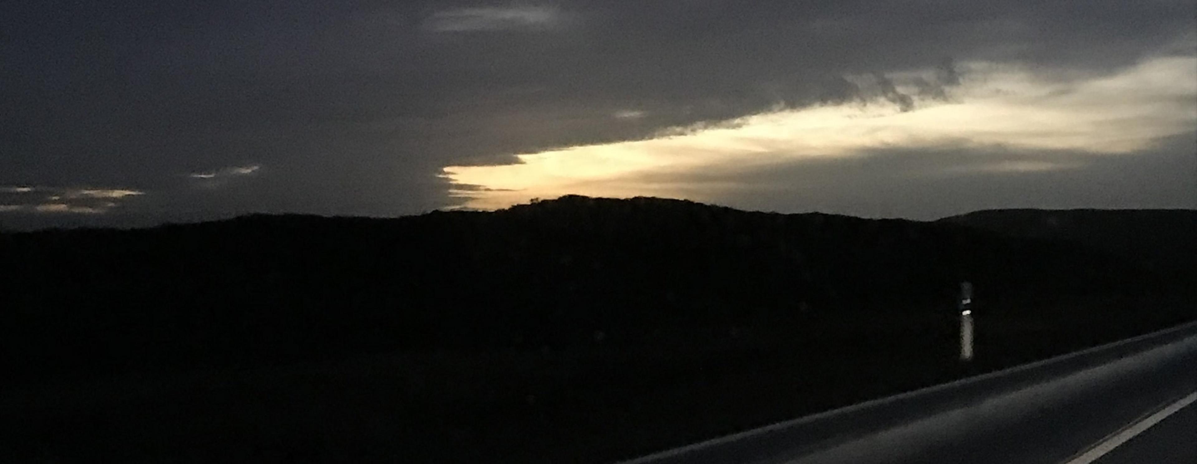 Sonnenaufgang hinter einer sehr dunklen Wolke, betrachtet aus einem Auto, das auf einer Landstraße fährt.
