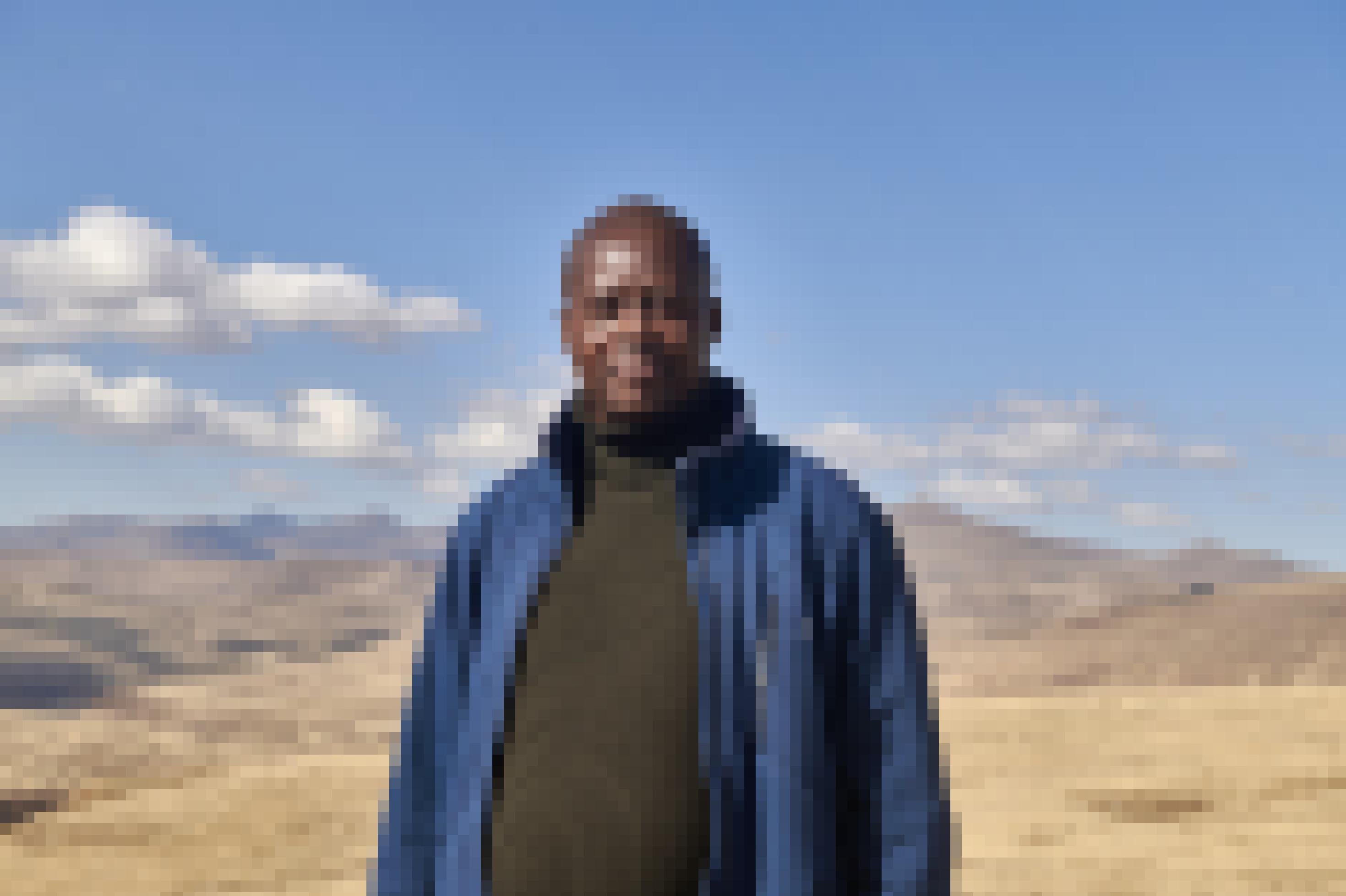 Seqhee steht vor der Berglandschaft, er trägt einen Rollkragen-Pullover, eine blaue Jacke und schaut in die Kamera