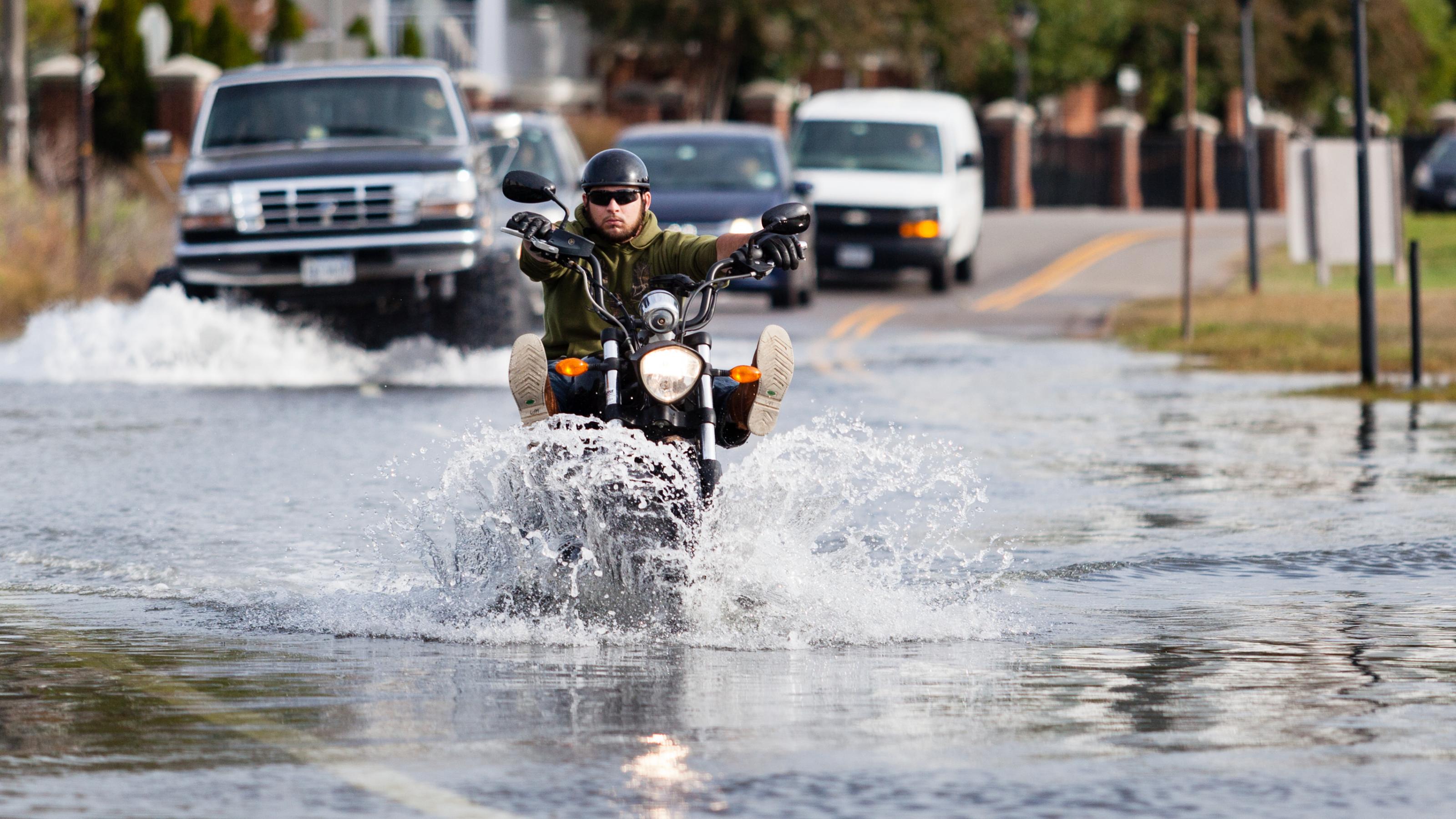 Füße hoch: Das Wasser steht in Norfolk/Virginia auf der Straße, und ein Motorrad-Fahrer fährt langsam durch das Wasser. Im Hintergrund warten Autos, unter anderem ein Geländewagen mit extra-großen Reifen. Foto: Will Parson/Chesapeake Bay Program