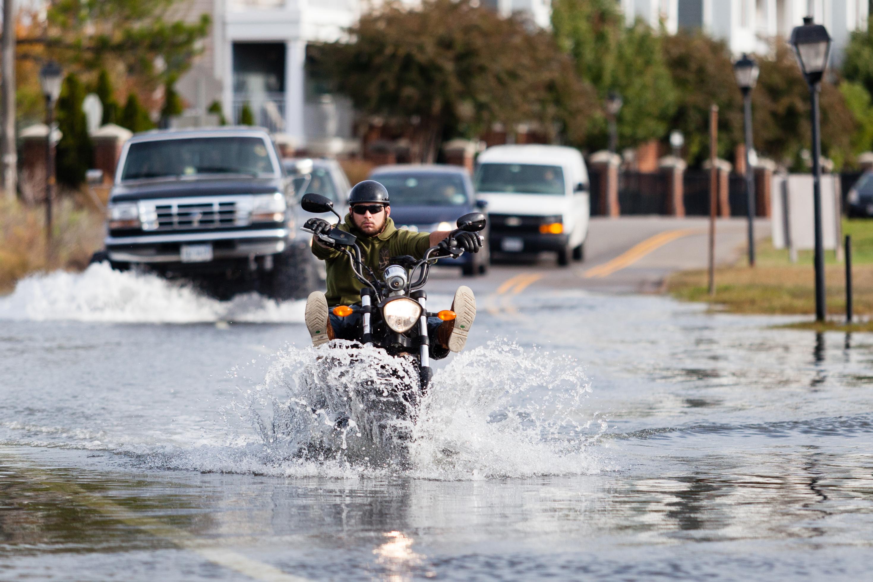 Füße hoch: Das Wasser steht in Norfolk/Virginia auf der Straße, und ein Motorrad-Fahrer fährt langsam durch das Wasser. Im Hintergrund warten Autos, unter anderem ein Geländewagen mit extra-großen Reifen. Foto: Will Parson/Chesapeake Bay Program