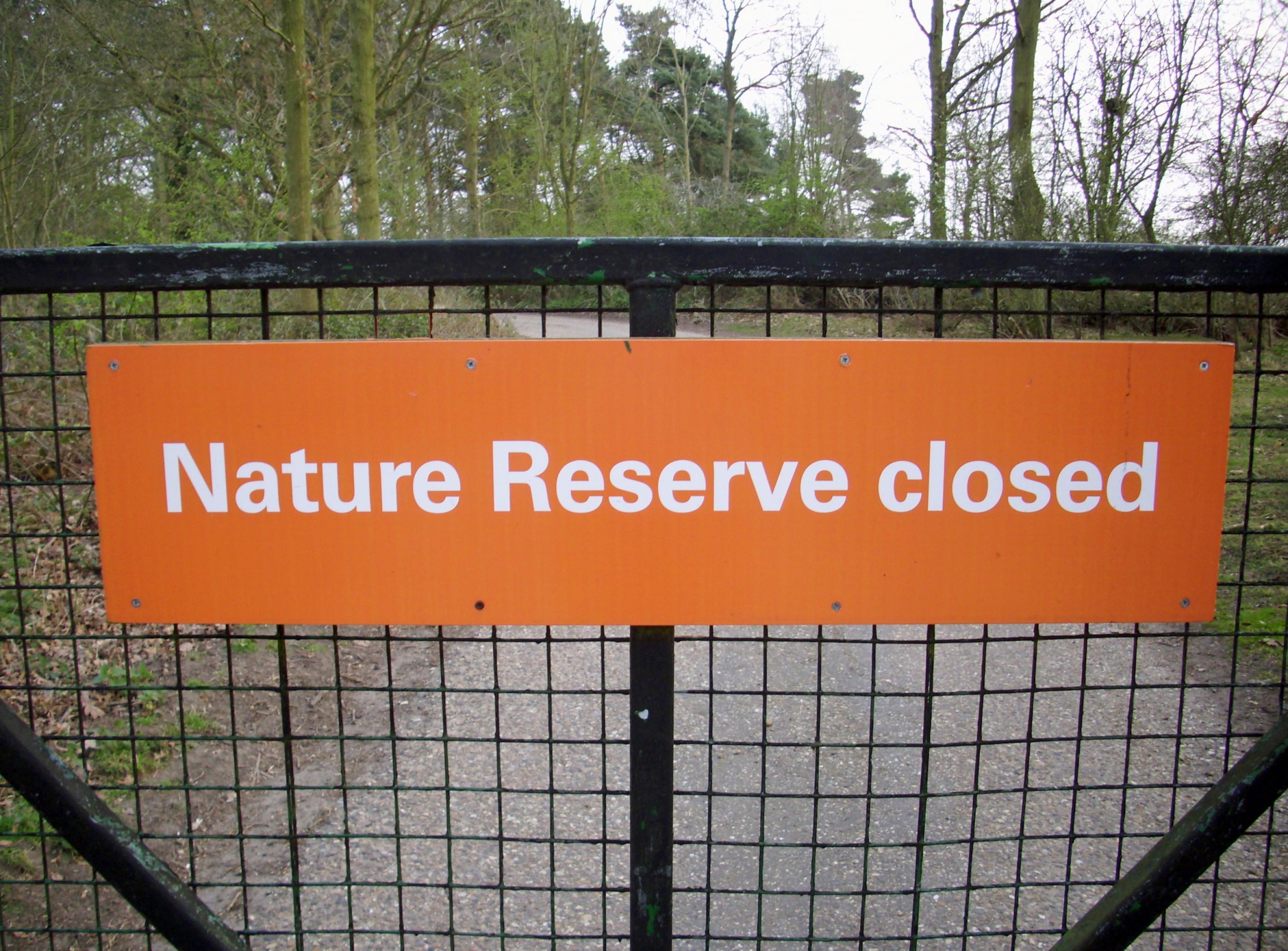 Das Bild zeigt ein Schild mit der Aufschrift „Nature Reserve Closed“ an einem Metallzaun vor einem Wald.