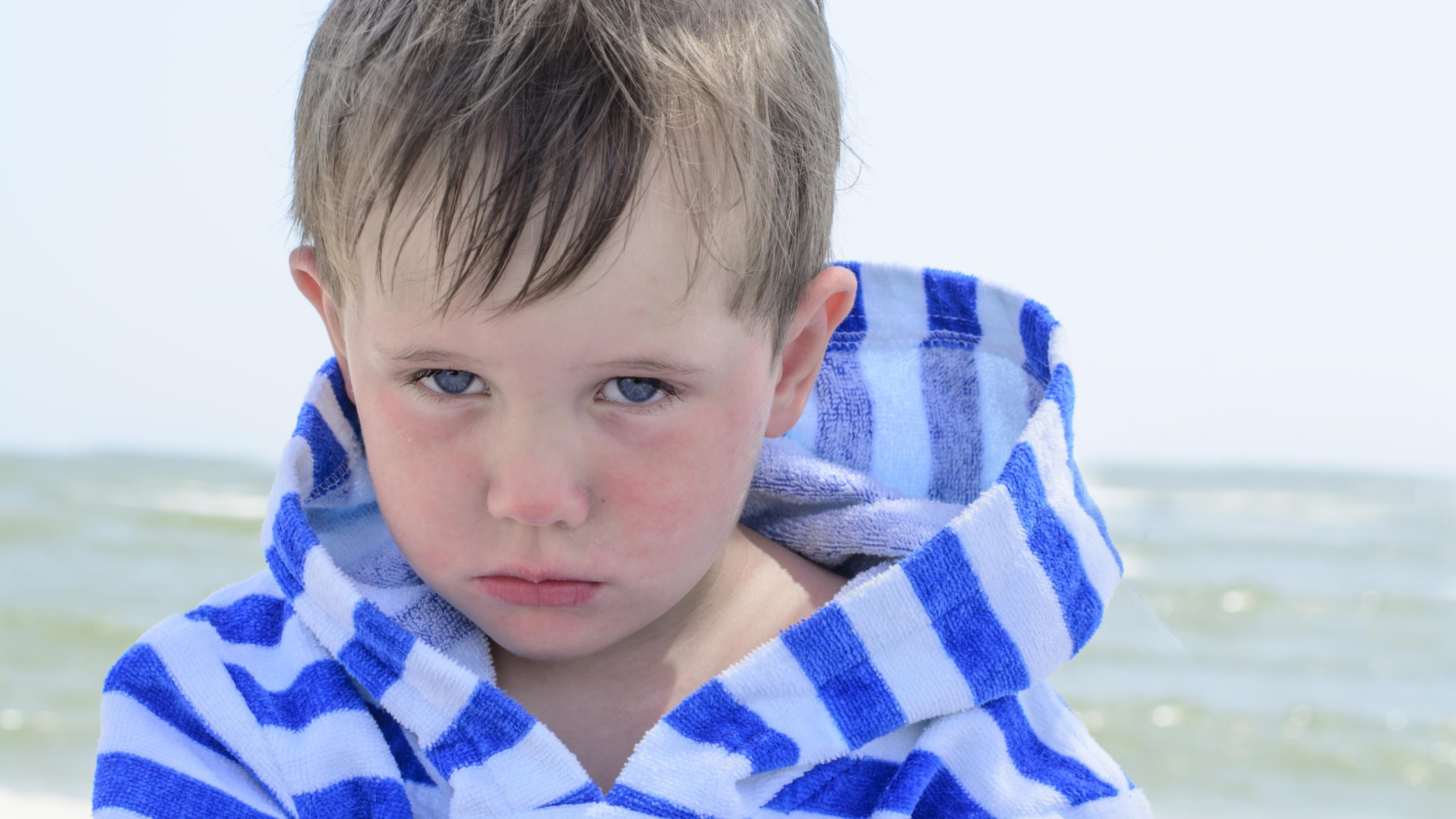 Ein Junge schaut traurig in die Kamera. Er trägt einen blau-weiß gestreiften Bademantel. Im Hintergrund ist das Meer zu sehen. Es ist ein sonniger Tag.