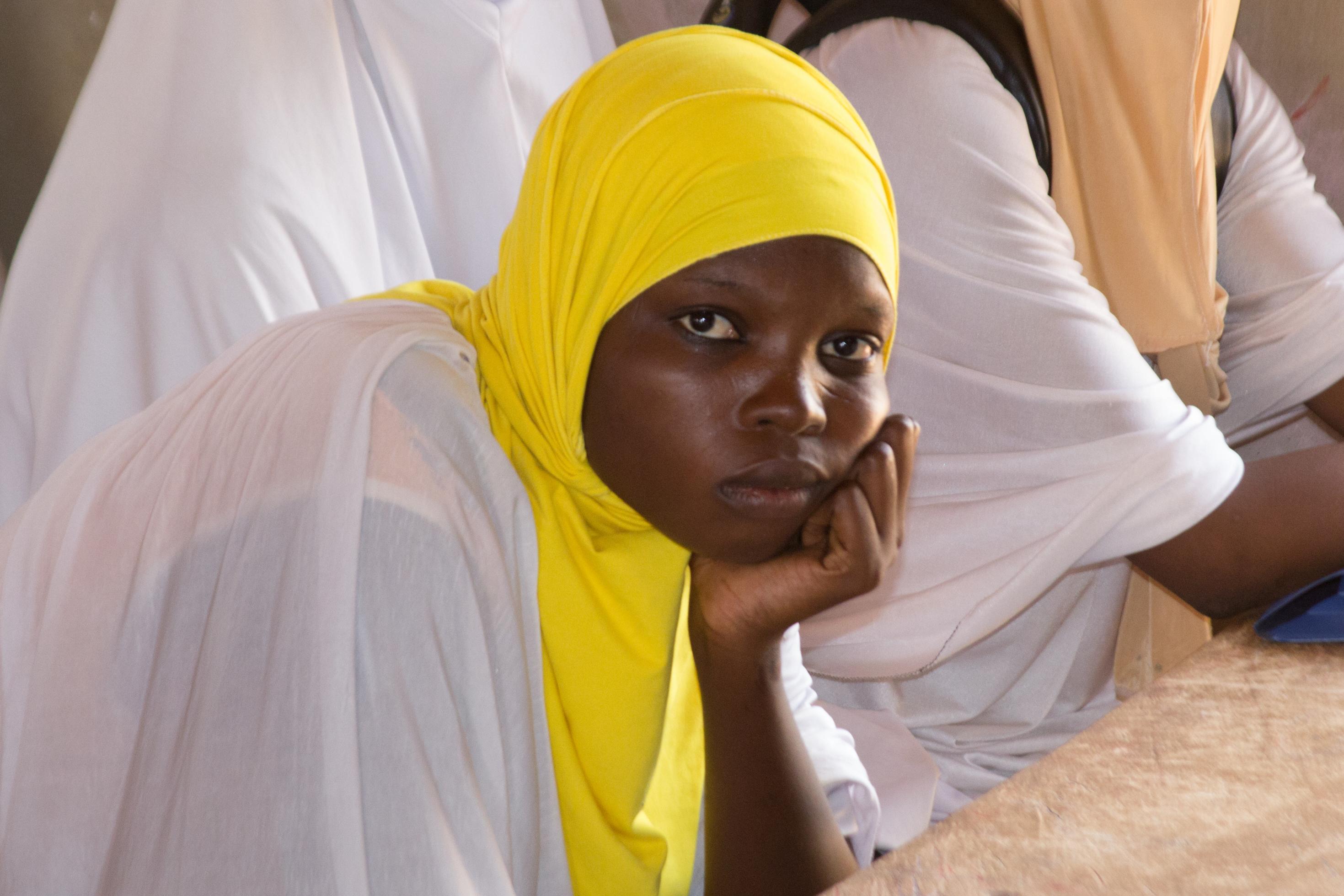 Eine Schülerin blickt die Betrachterin/ den Betrachter direkt an, um sie herum sind weitere Schülerinnen zu sehen. Alle tragen weiße Kleidung und farbige Kopftücher.