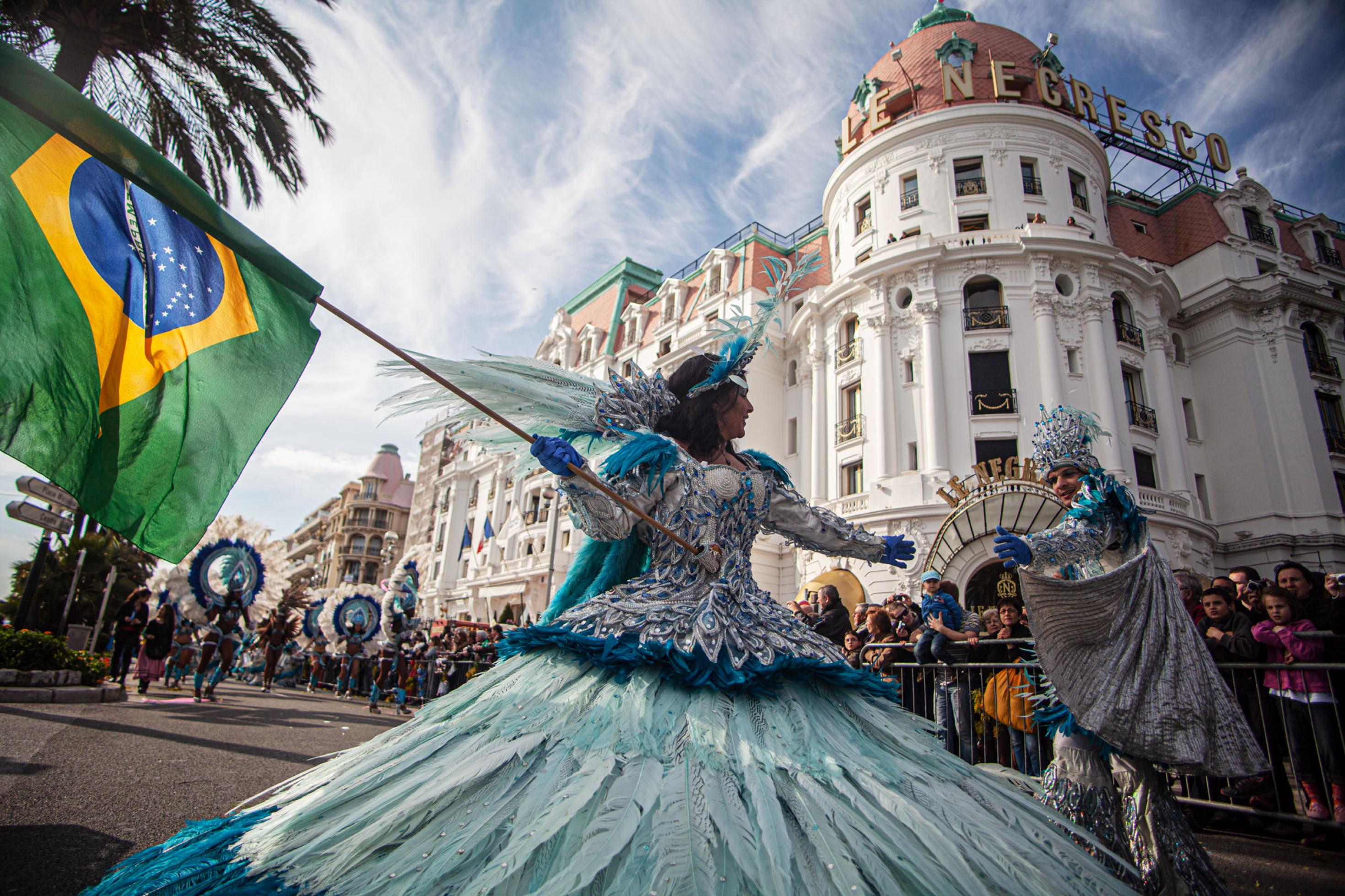 Bunt gekleidete Tänzer und Tänzerinnen beim Karneval in Nizza vor dem Hotel Negresco mit brasilianischer Flagge