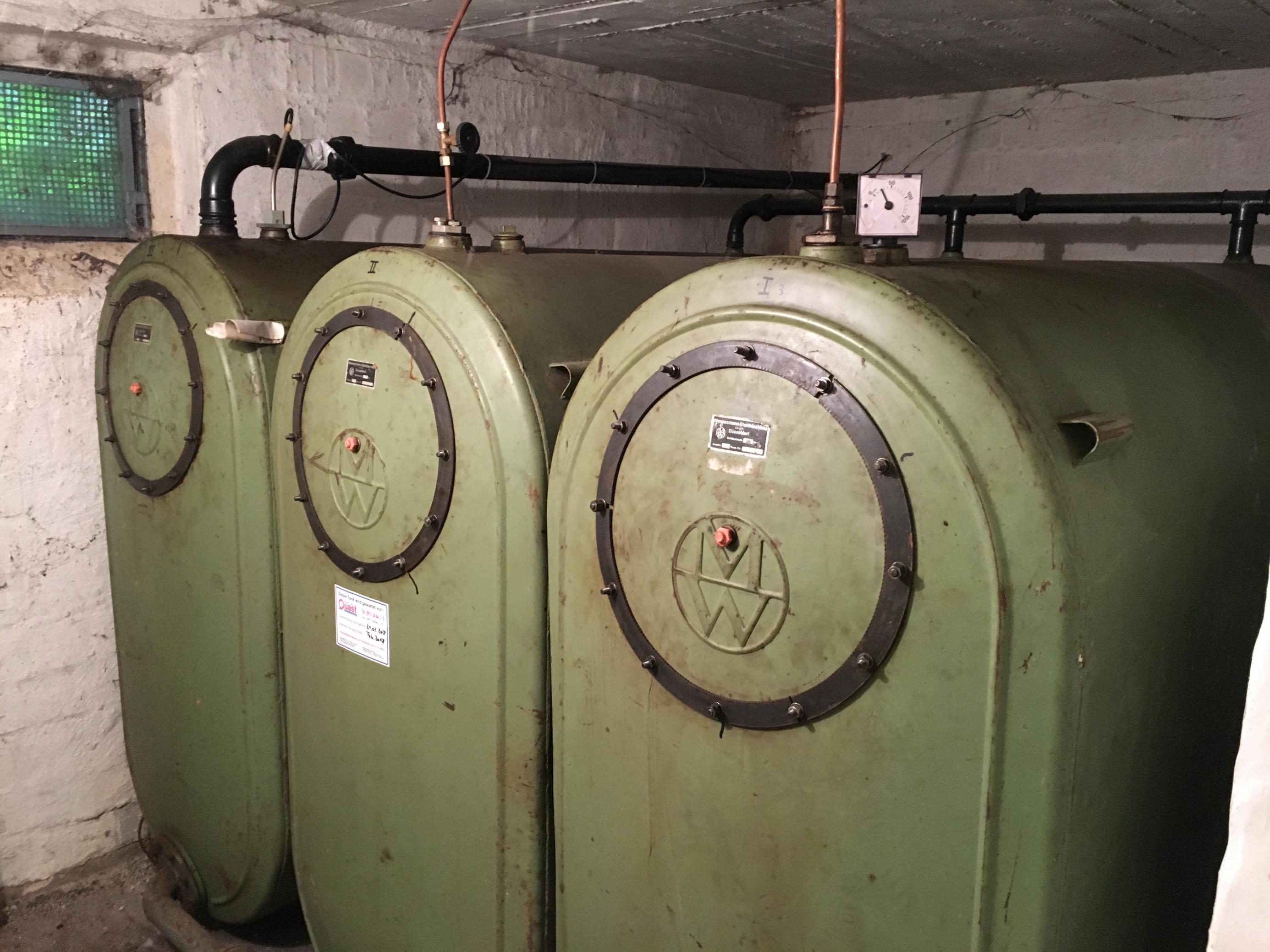 Ein dreifacher Öltank auf oliv-grünem Stahl in einem Kellerraum. Längst sind solche Anlagen ein Sanierungsfall. In Zukunft könnte eine solche Anlage durch eine Wärmepumpe ersetzt werden, dann lassen sich Wärme und Duschwasser mit Strom aus erneuerbaren Quellen erzeugen.