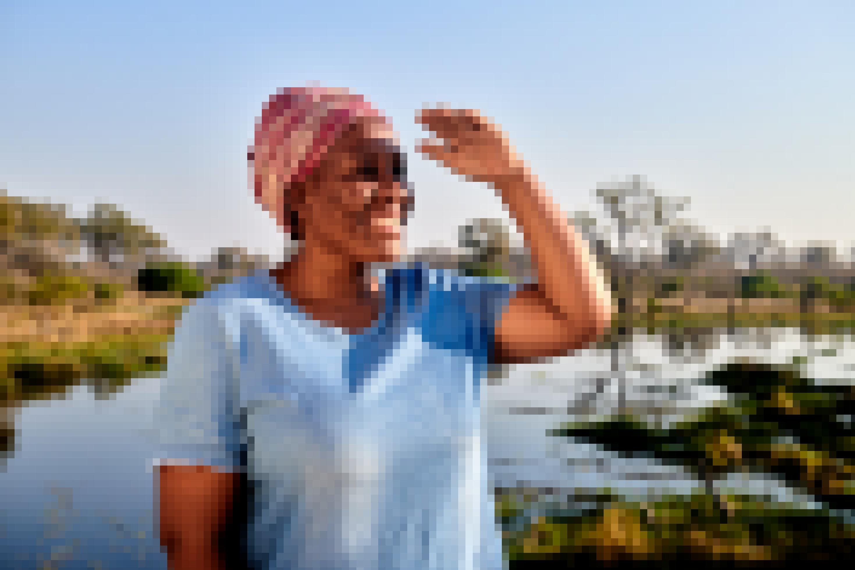 Die Frau schirmt ihre Augen mit einer Hand vor dem Sonnenlicht ab und schaut lächelnd in die Ferne, sie trägt ein rosa-gemustertes Tuch auf dem Kopf und ein blaues Kleid