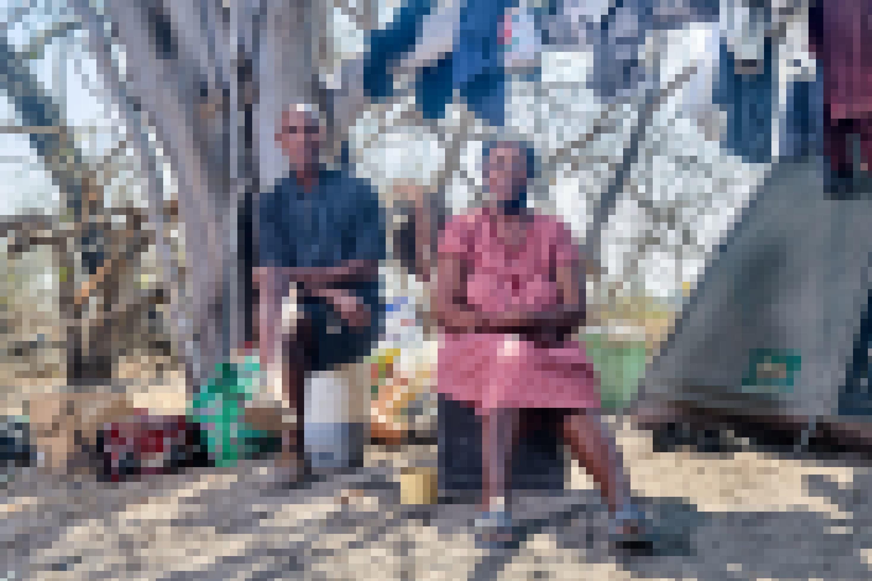Das Paar sitzt auf zwei Plastikcontainern vor einem Baum und einer Wäscheleine in ihrem Zeltlager. Er trägt kurze Hosen, sie ein rot-weiß-kariertes Kleid