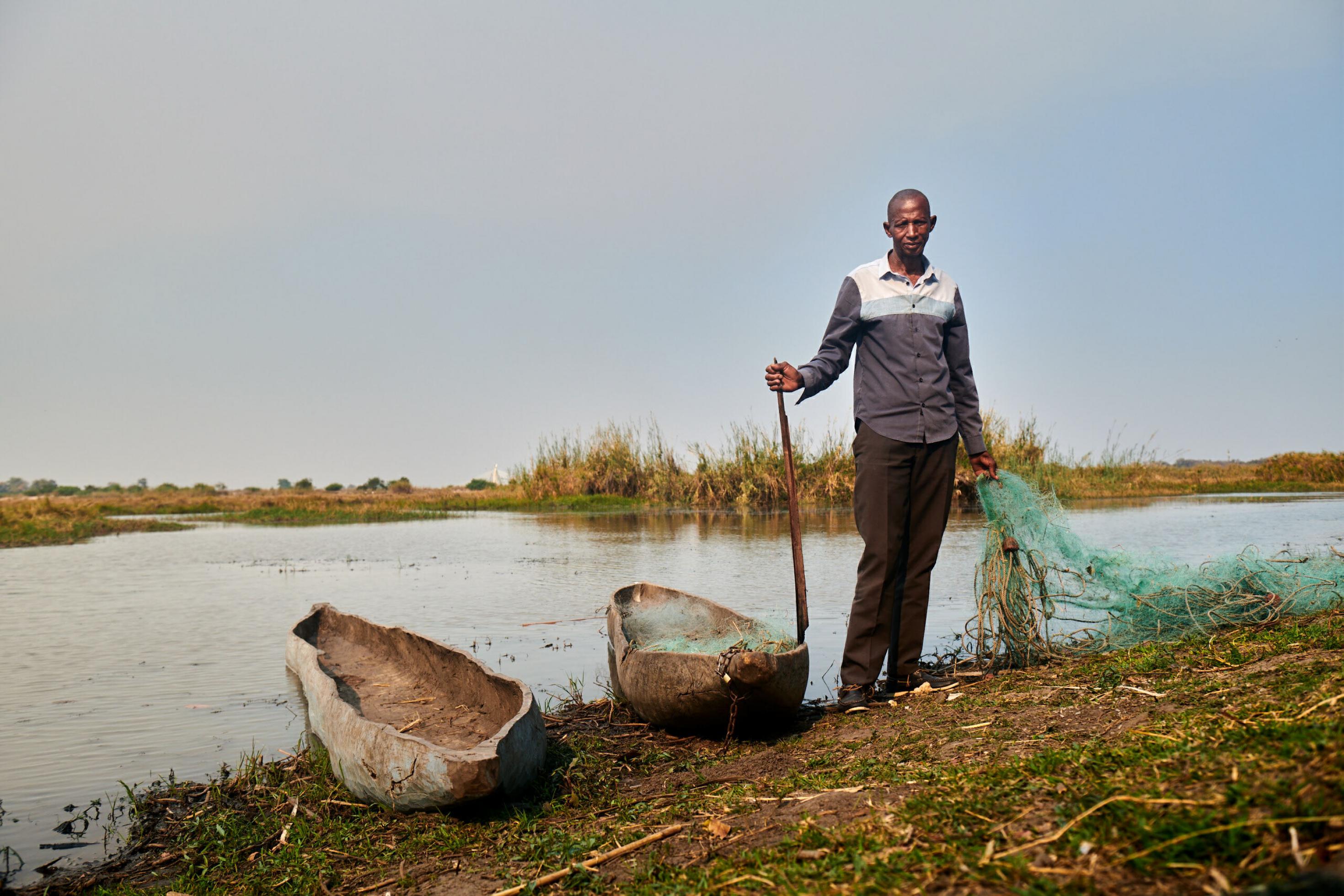 Der Fischer steht mit einem Netz in der Hand neben zwei Holzbooten am Ufer des Okavango