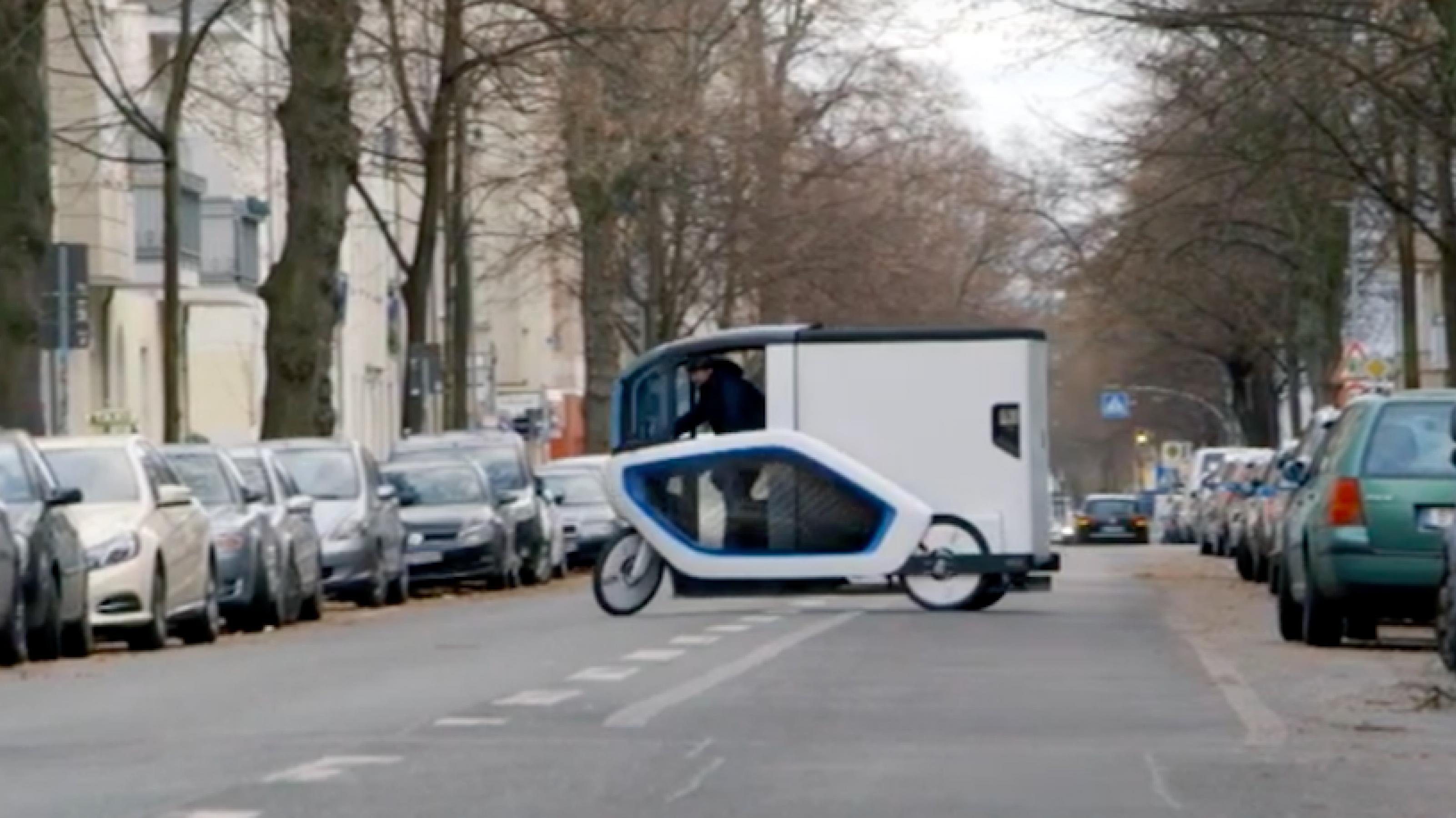 Die Ono ist ein Schwertransportern unter den Lastenrädern. Bei dem Wendemanöver mitten auf der Fahrbahn ist die Silhouette des weißen Gefährts mit der Fahrerkabine gut erkennbar. Mit ihrer Höhe von rund zwei Meter überragt sie die rechts und links parkenden Autos.