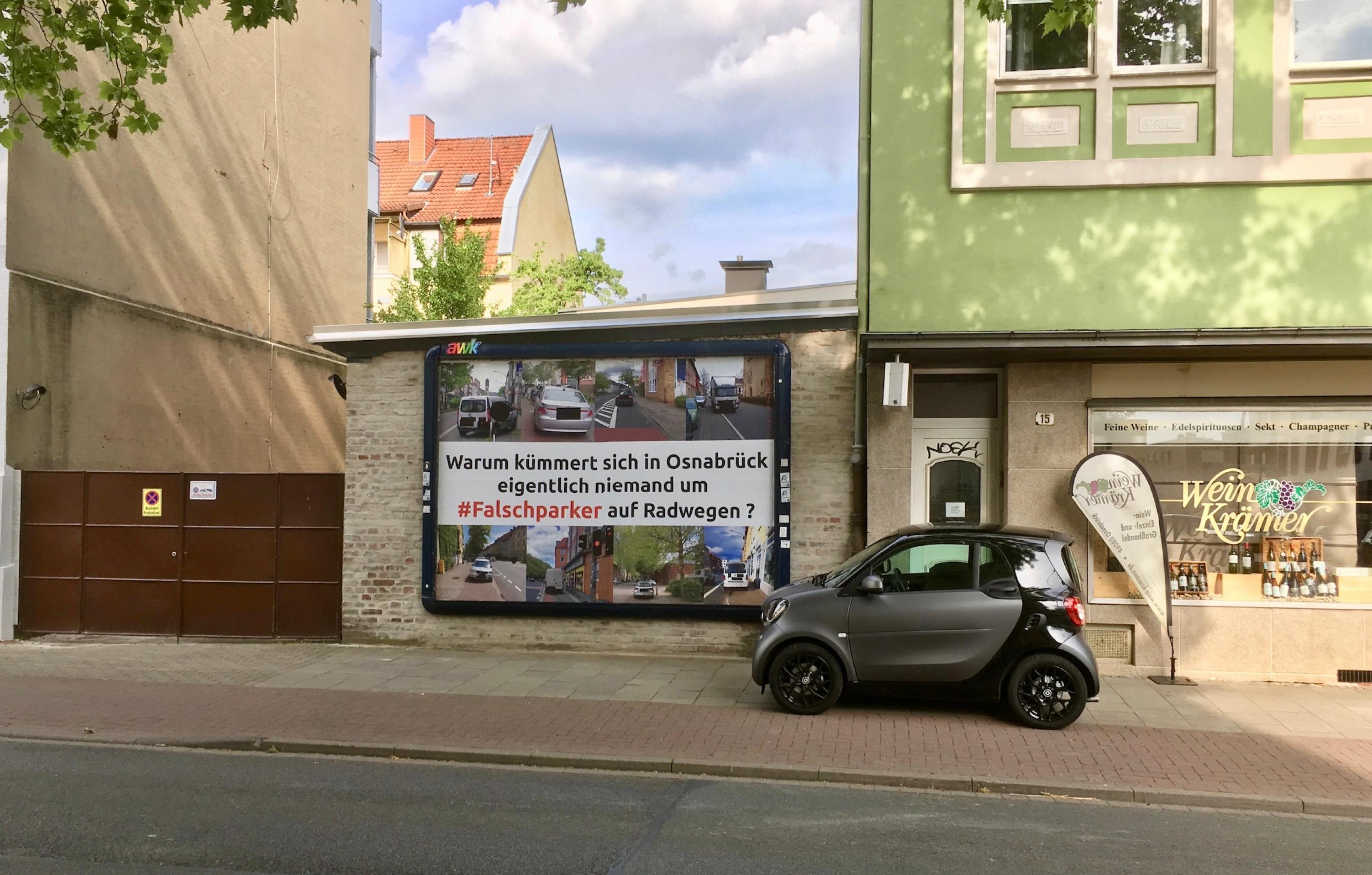 Ein grau-schwarzfarbener Smart steht auf den Gehweg neben einem Radweg. Er steht direkt neben einer Plakatwerbung die Falschparker mit der Aufschrift:" Warum kümmert sich in Osnabrück eigentlich niemand um #Falschparker auf Radwegen?"