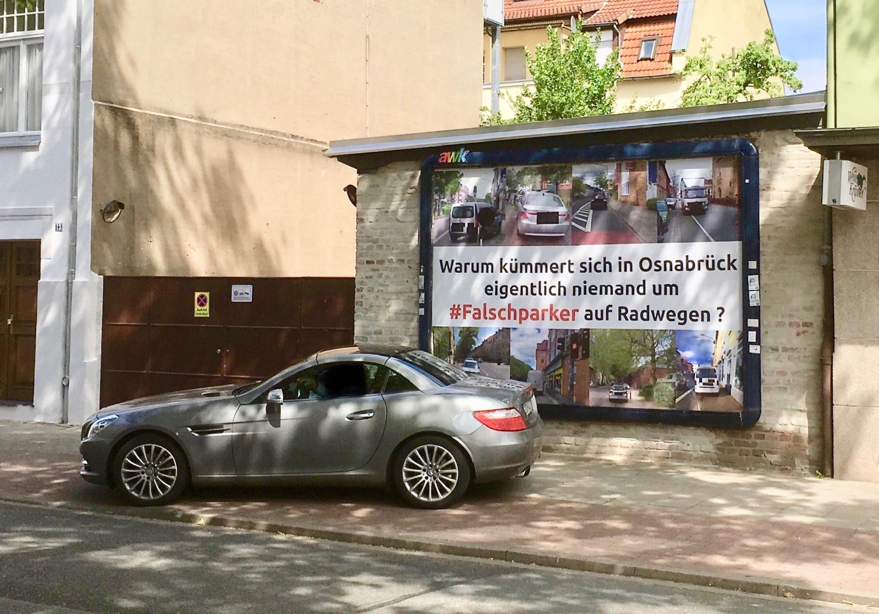 Ein Sportwagen von BMW steht quer auf dem Rad und Fußweg. Es steht mit dem Heck vor dem Plakat mit dem Schriftzug „Warum kümmert sich in Osnabrück eigentlich niemand um #Falschparker auf Radwegen?“