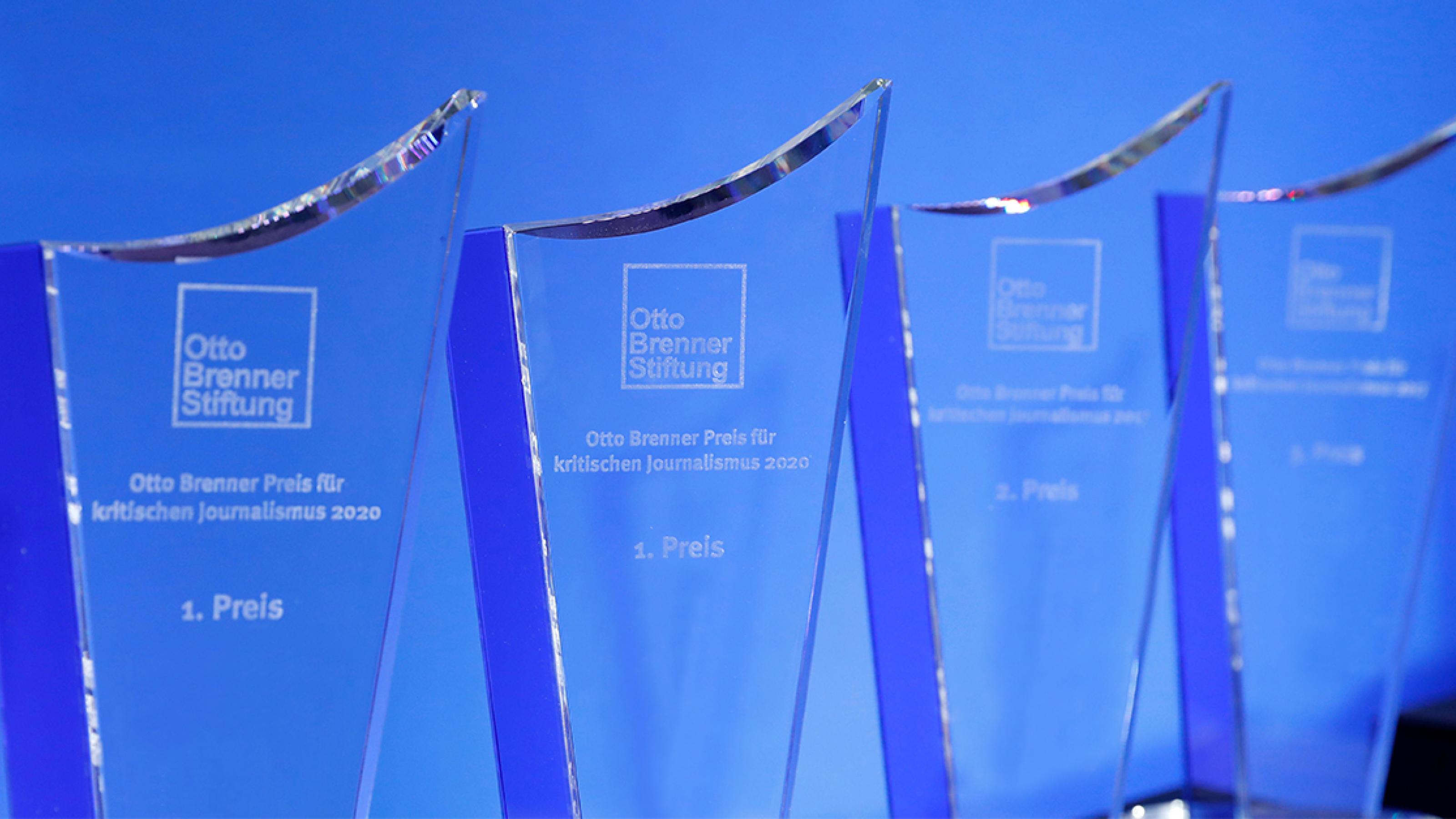 Mehrere Otto-Brenner-Preis-Trophäen aus Glas stehen vor einem blauen Hintergrund.