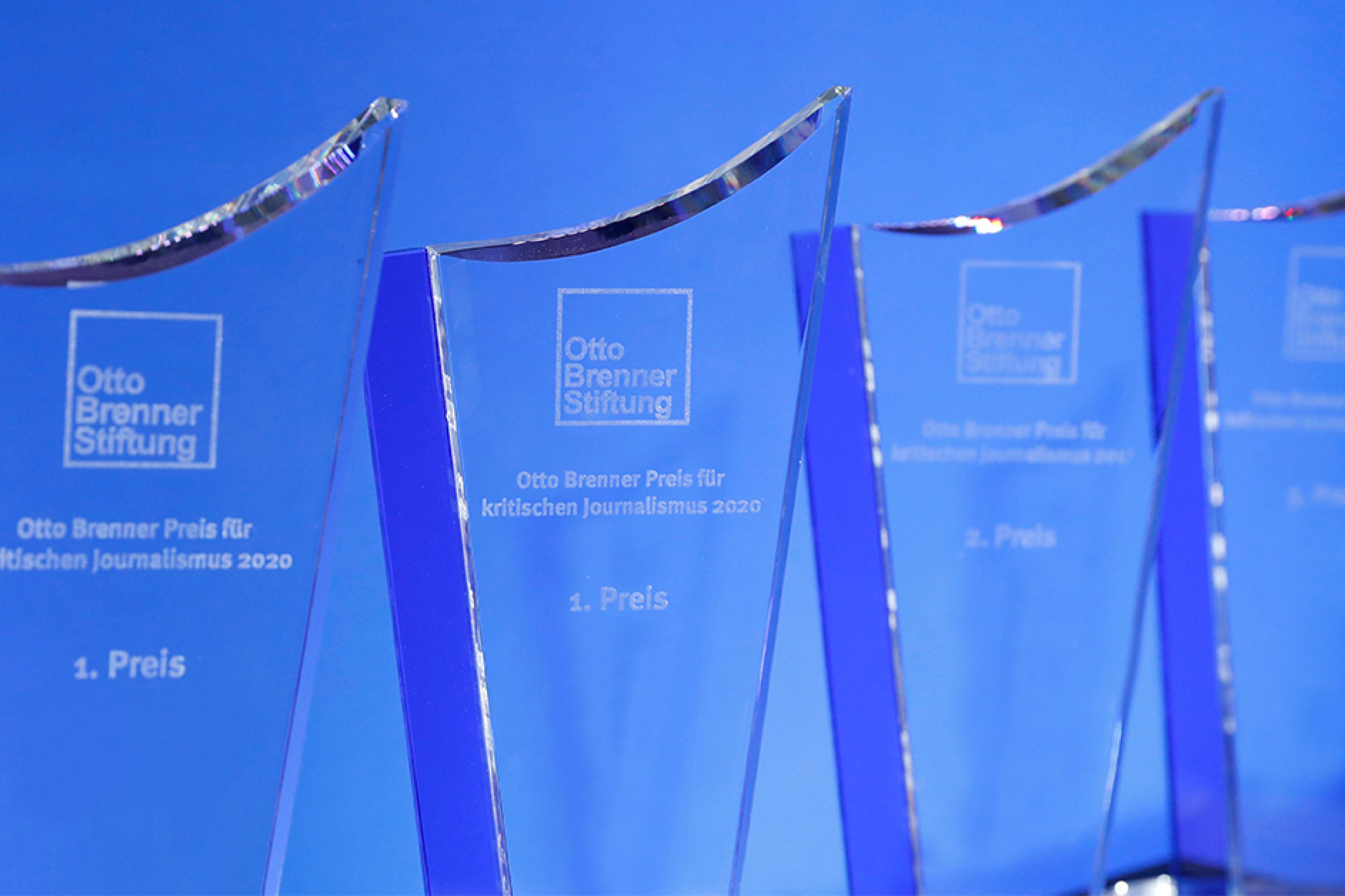 Mehrere Otto-Brenner-Preis-Trophäen aus Glas stehen vor einem blauen Hintergrund.