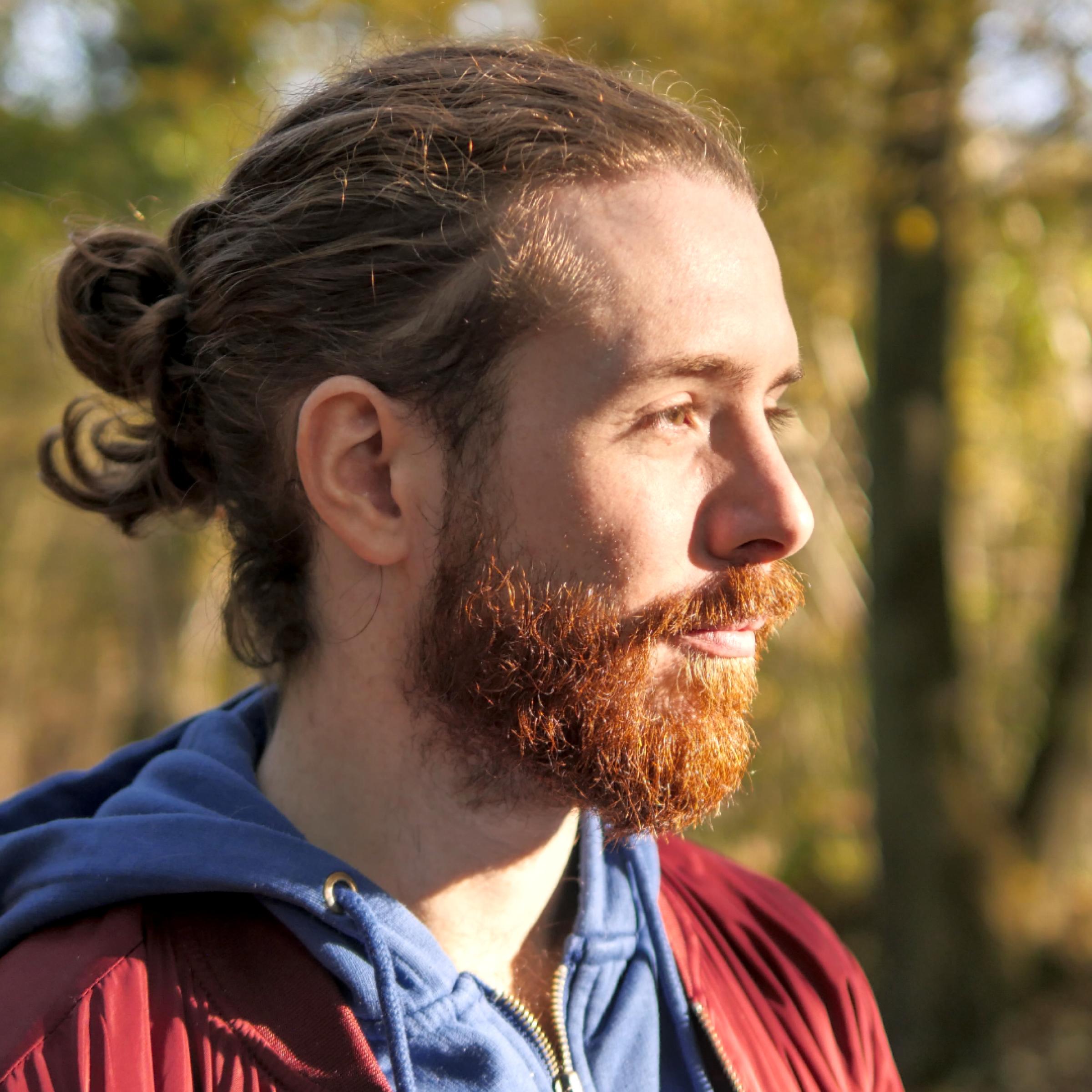 Auf dem Bild sieht man den Kopf eines rothaarigen Mannes mit Bart und langen, gebundenen Haaren aus der Profil-Perspektive. Er steht vor dem Hintergrund eines kleinen Waldes.
