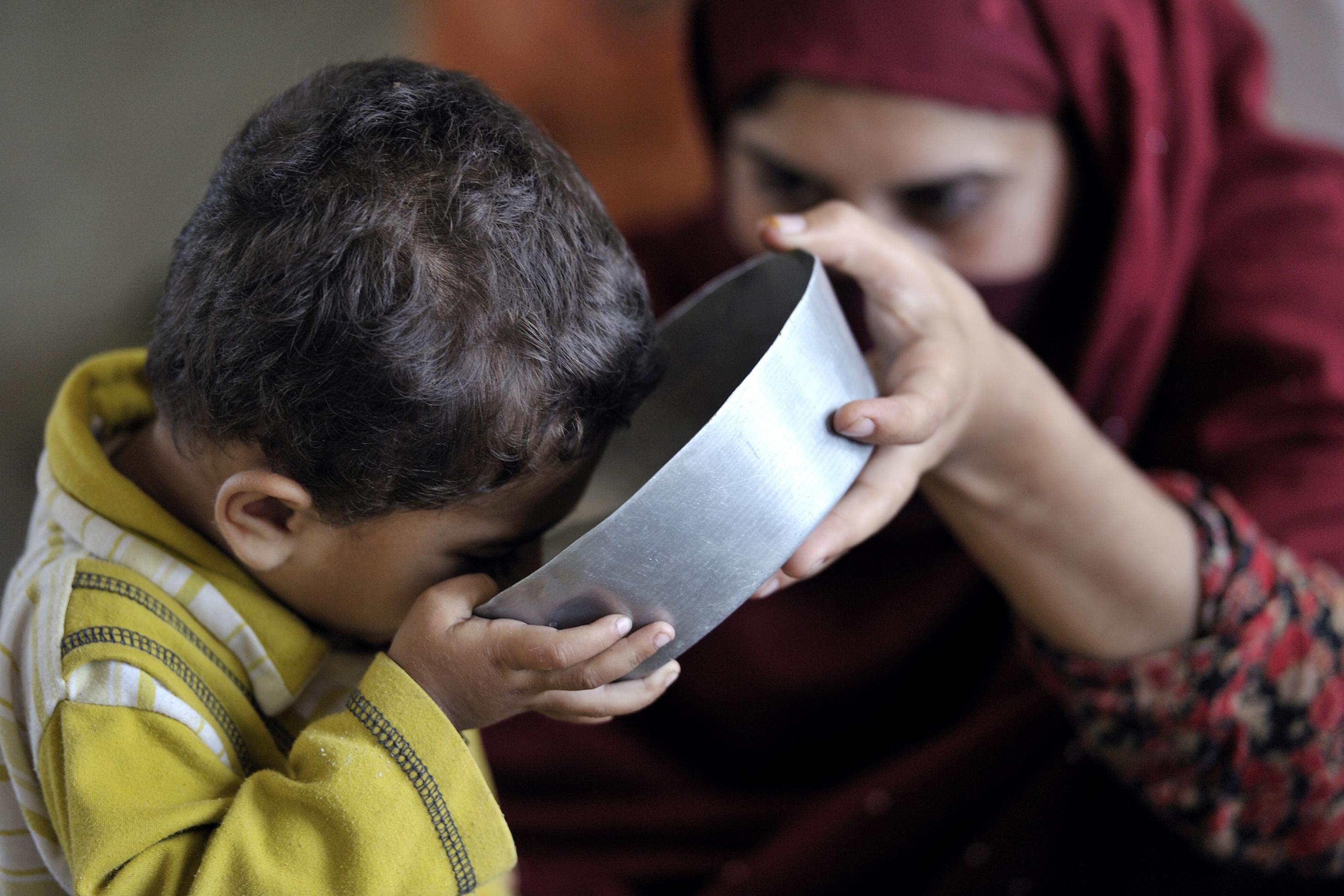Ein kleines Kind trinkt aus einer Metallschüssel, im Hintergrund ist seine Mutter zu sehen, die das Gefäß hält. Viele Bewohner von überfluteten Orten in Pakistan mussten fliehen und fanden Schutz in Lagern und Versorgung von internationalen Unterstützern. Hier trinkt ein durstiges Kind sauberes Wasser, das UNICEF-Helfer bereitgestellt haben.