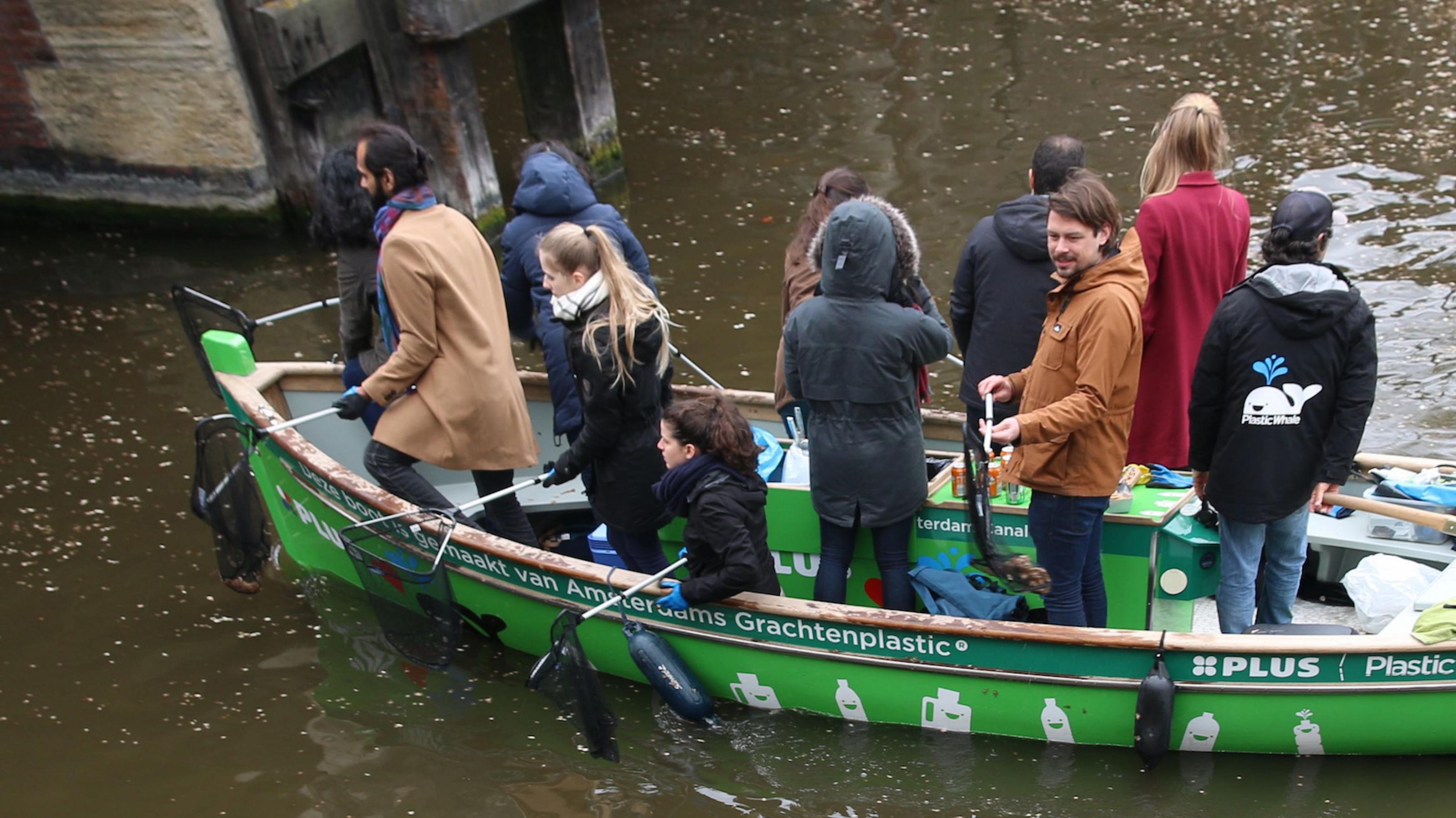 Ein grünes Boot auf einer Gracht in Amsterdam, darin stehen und sitzen elf junge Leute. Sie haben Käscher in der Hand. Die Aufschrift des Bootes besagt, dass die Mitfahrer Plastikmüll aus dem Wasser fischen. Jeder Fang wird laut bejubelt.