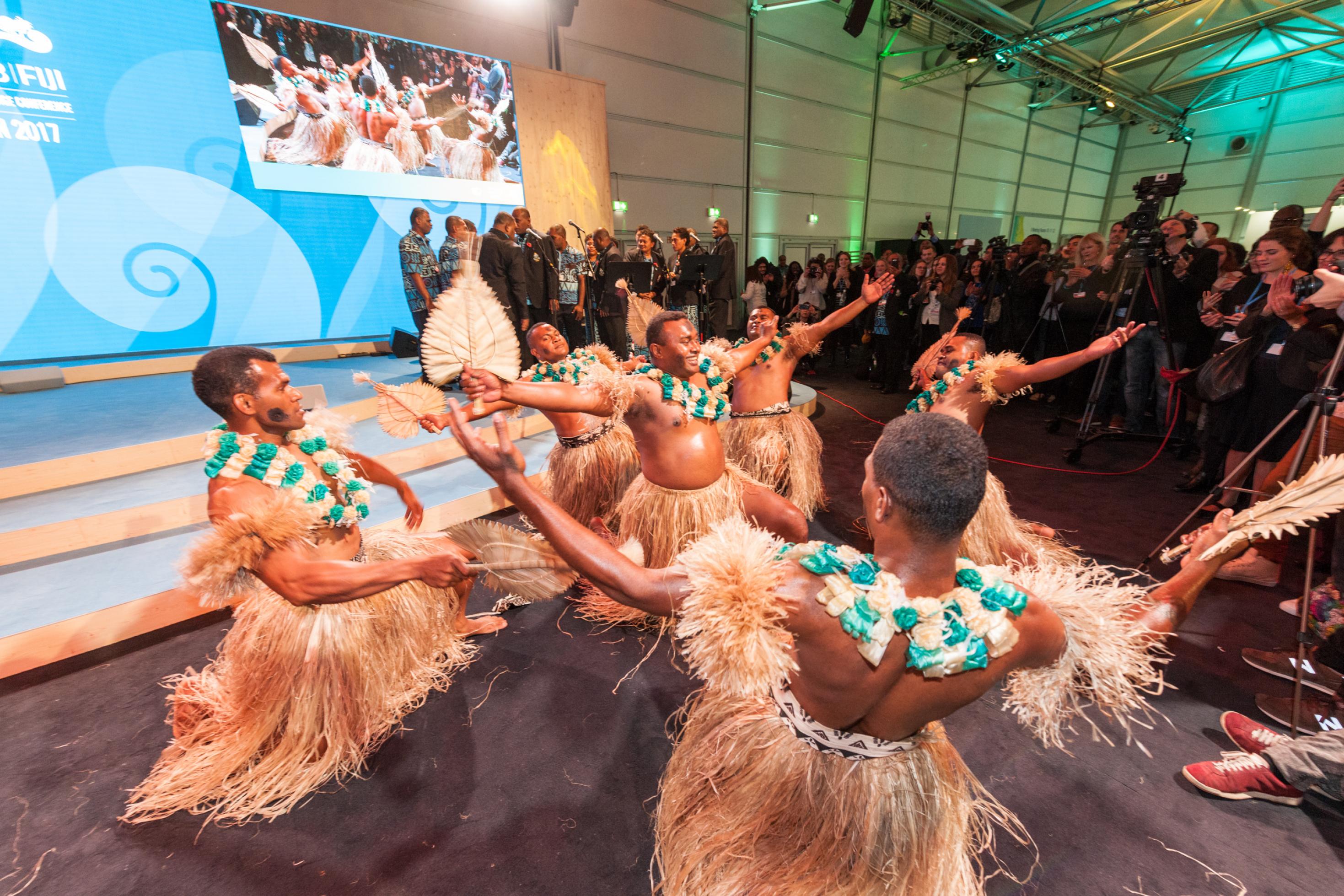 Eine Gruppe von Männern mit Baströcken, nackten Oberkörpern und blau-weißen Blütenkränzen am Hals dreht sich mit halbhoch erhobenen Armen im Kreis: Polynesische Tänzer bei einem Empfang während der COP in Bonn