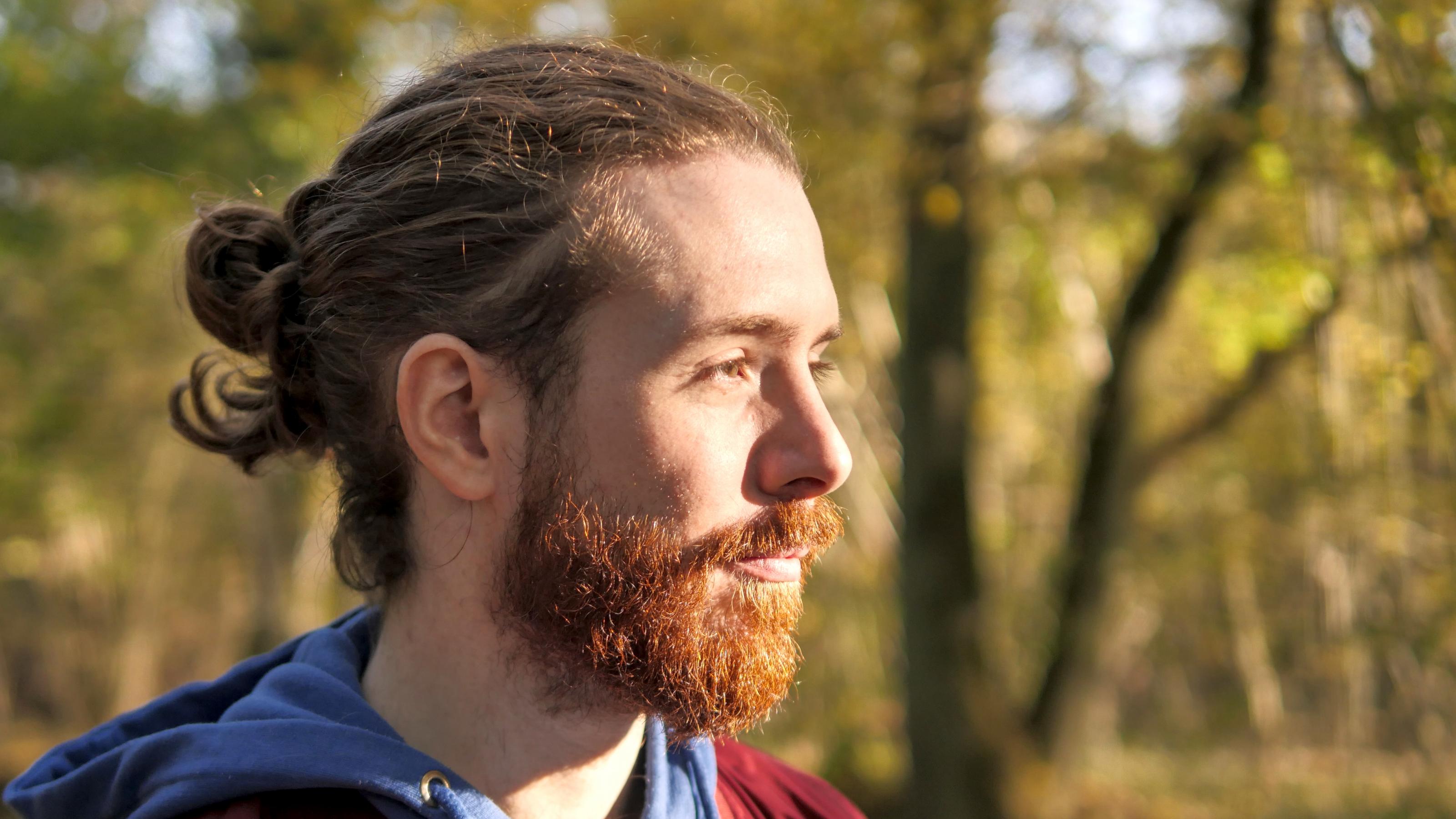 Auf dem Bild sieht man den Kopf eines rothaarigen Mannes mit Bart und langen, gebundenen Haaren aus der Profil-Perspektive. Er steht vor dem Hintergrund eines kleinen Waldes.