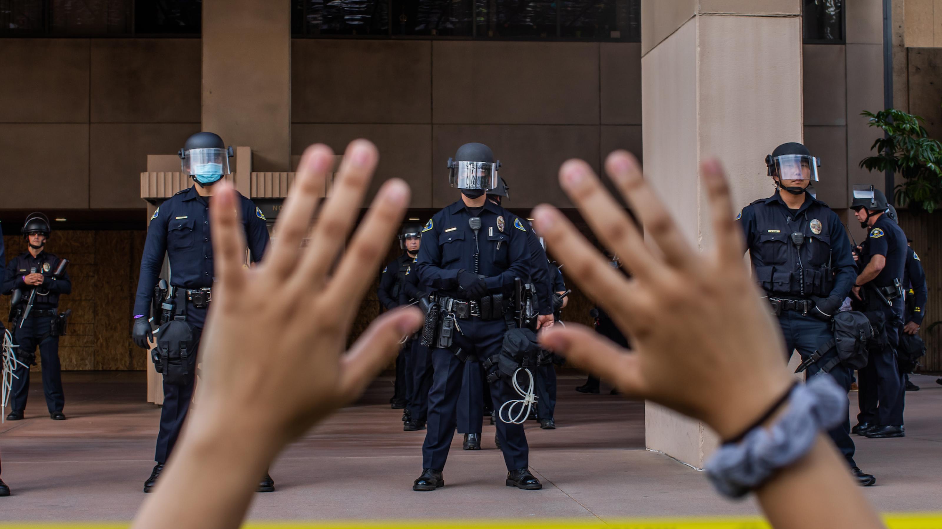 Ein Demonstrant hält seine Hände nach oben, während im Hintergrund Polizisten in Schutzausrüstung zu sehen sind.