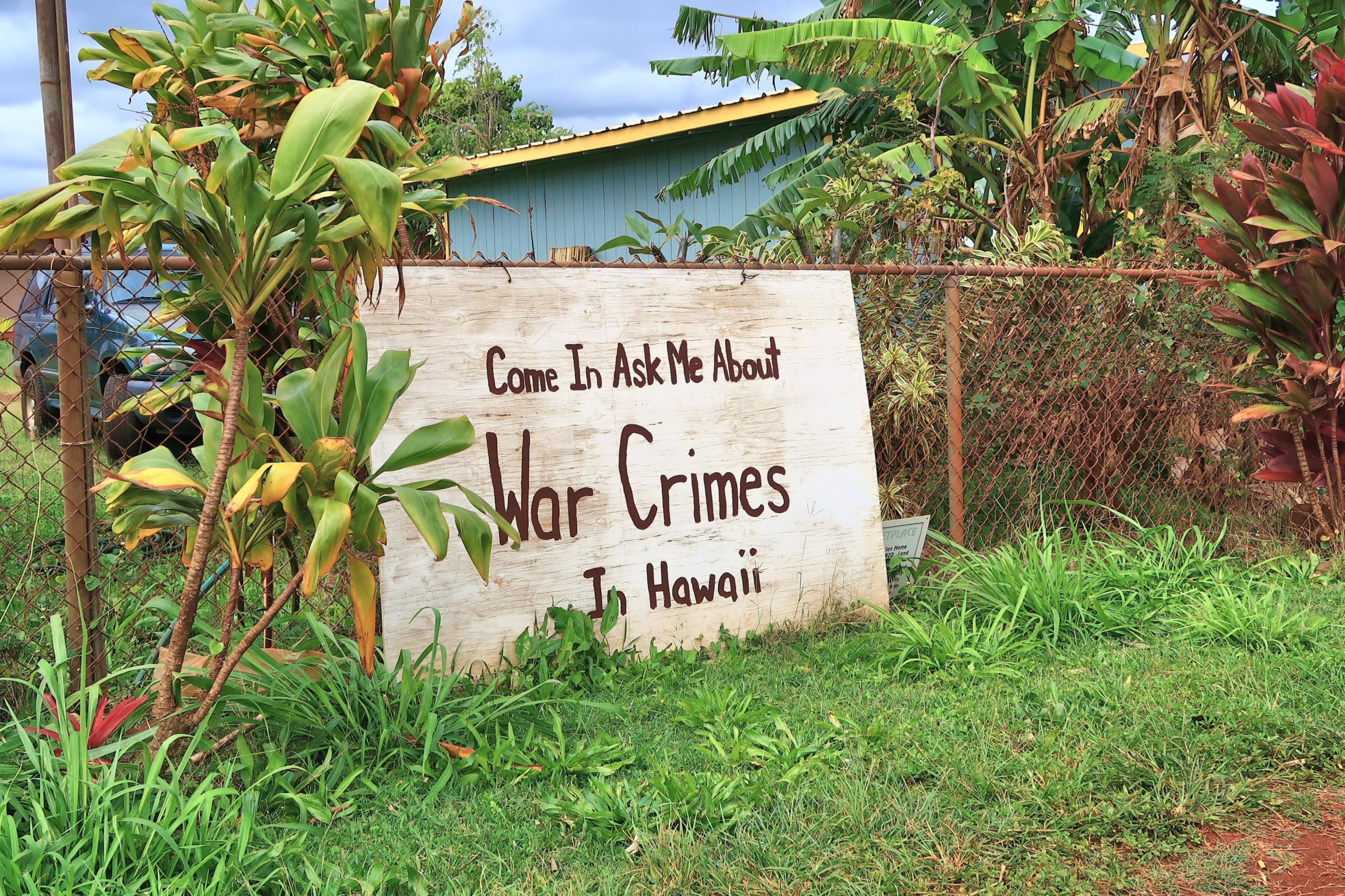 Ein Schild lehnt an einem Zaun, Aufschrift: „Come in and ask me about war crimes in Hawaii“