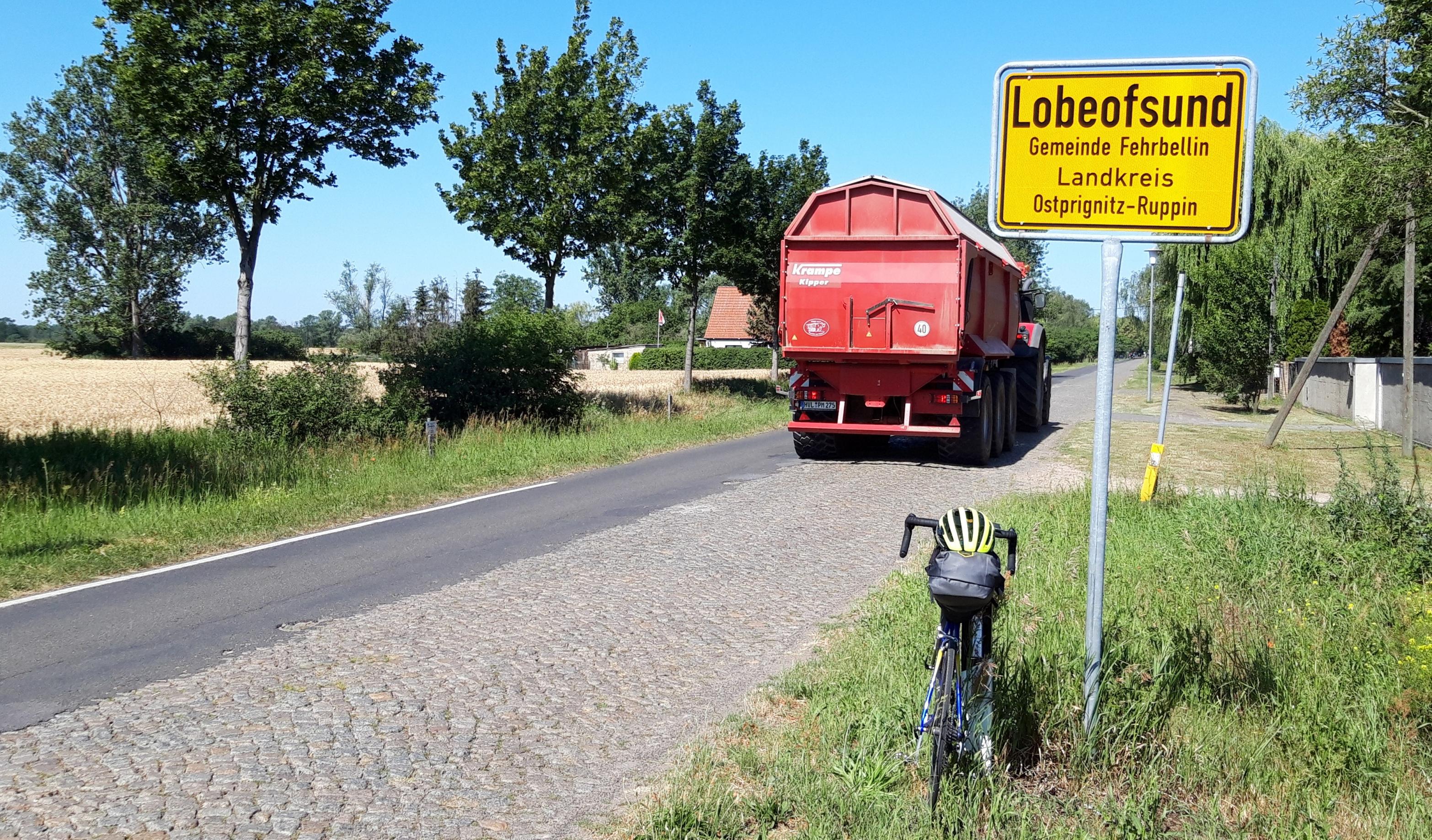 Das Rad lehnt am Ortsschild von Lobeofsund, auf der Kopfsteinpflasterstraße ist nur eine Spur asfaltiert.