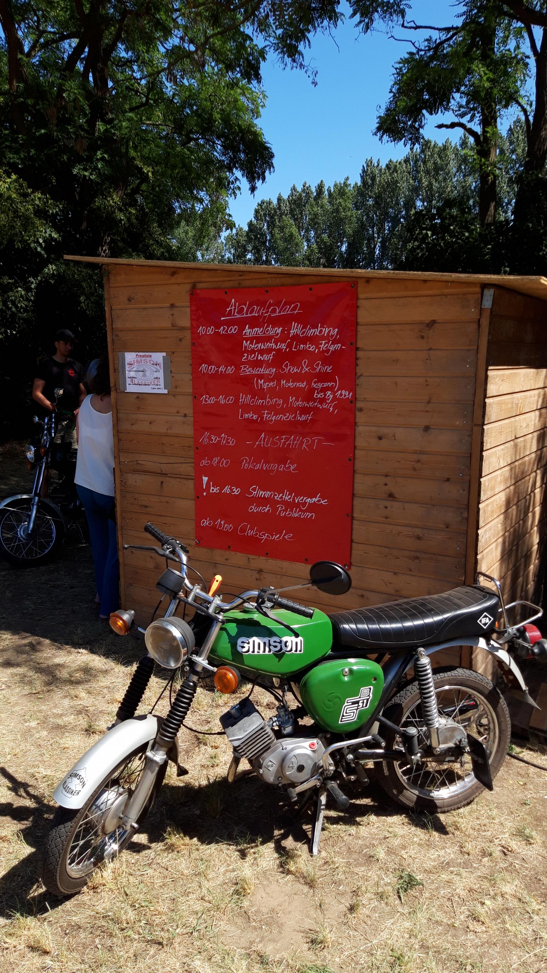 Per Hand geschrieben stehen die Veranstaltungsdetails auf einer großen Tafel, davor ein Motorrad