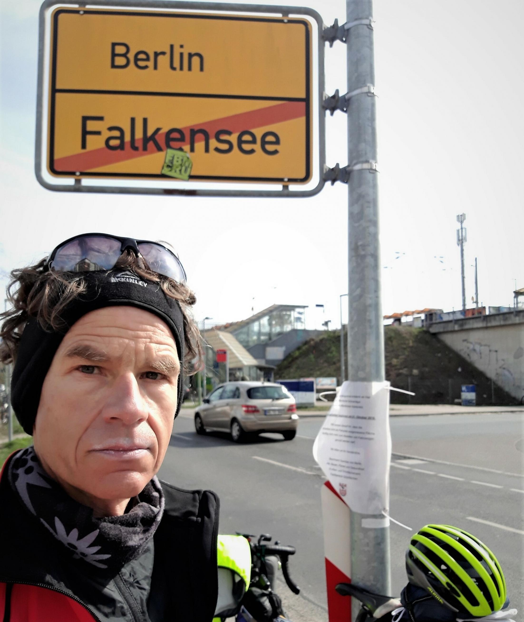 Ein abgekämpft wirkender Martin C. Roos macht ein Selfie am Ortsschild von Berlin, an dessen Pfahl ein offiziell wirkender Aushang befestigt ist. Im Hintergrund zu sehen: Der Bahnhof Albrechtshof.