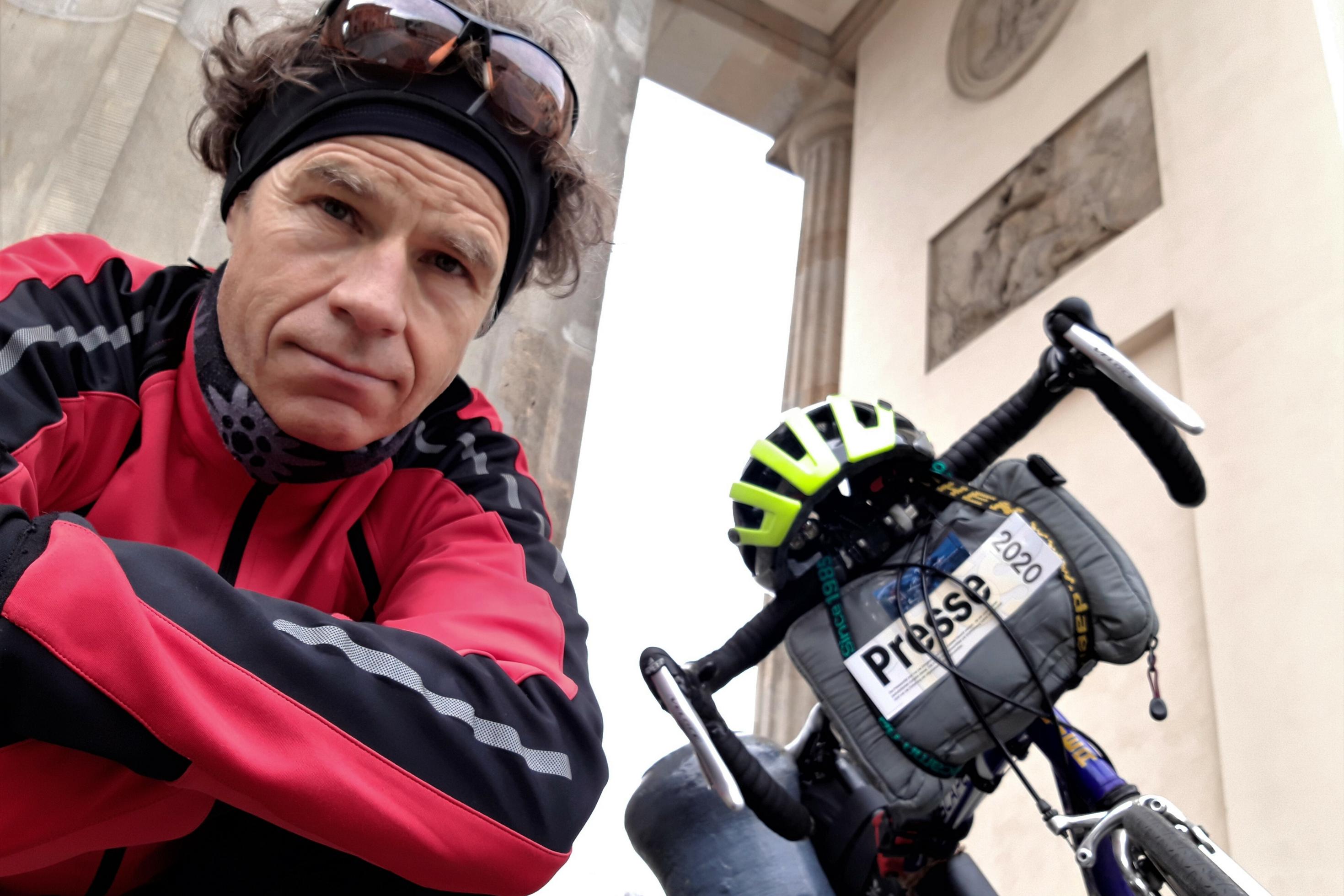 Martin C. Roos schaut in die Kamera, im Hintergrund ein Teil seines Rads und des Brandenburger Tors.
