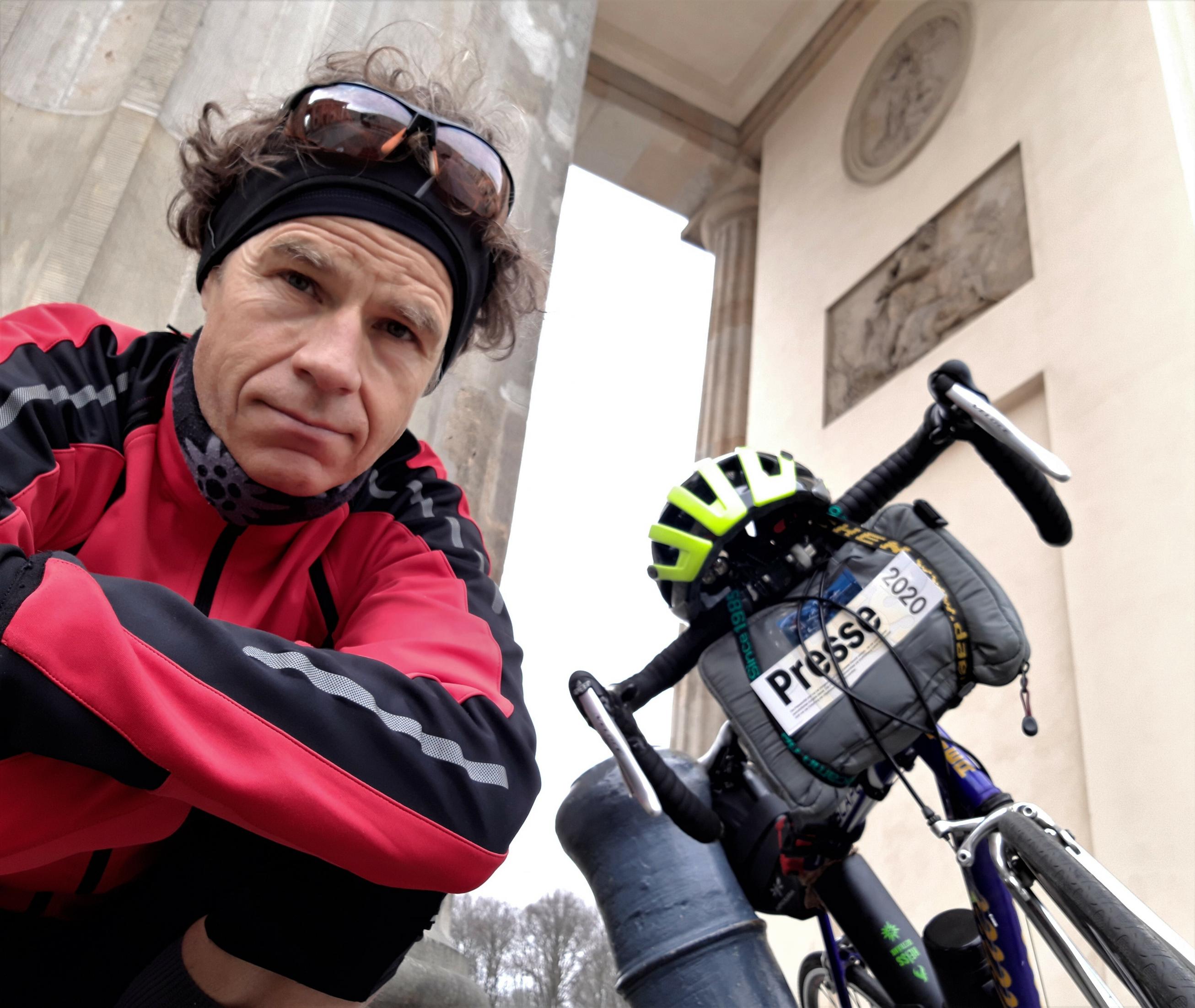 Martin C. Roos schaut in die Kamera, im Hintergrund ein Teil seines Rads und des Brandenburger Tors.