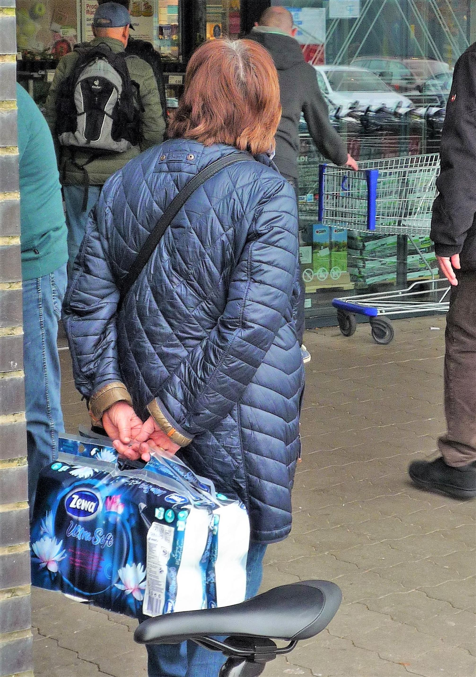Verschämt hält eine ältere Frau die zwei Riesenpackungen Toilettenpapier hinter dem Rücken, während sie am Supermarkteingang auf jemanden oder etwas wartet.