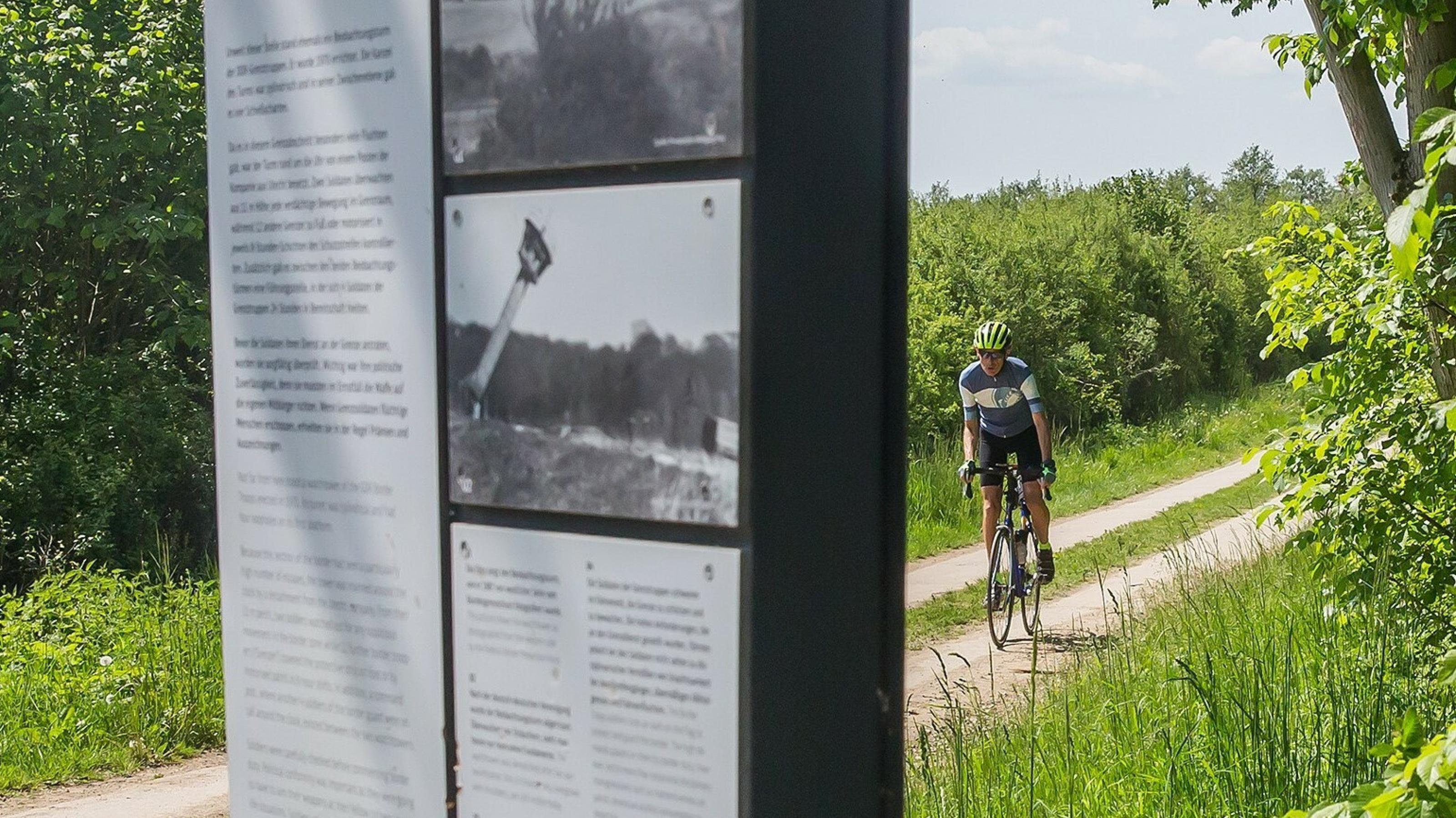 Martin C Roos fährt mit dem Rennrad auf einem alten Kolonnenweg, an dem – im Bildvordergrund – eine Schautafel des Museums zu sehen ist.