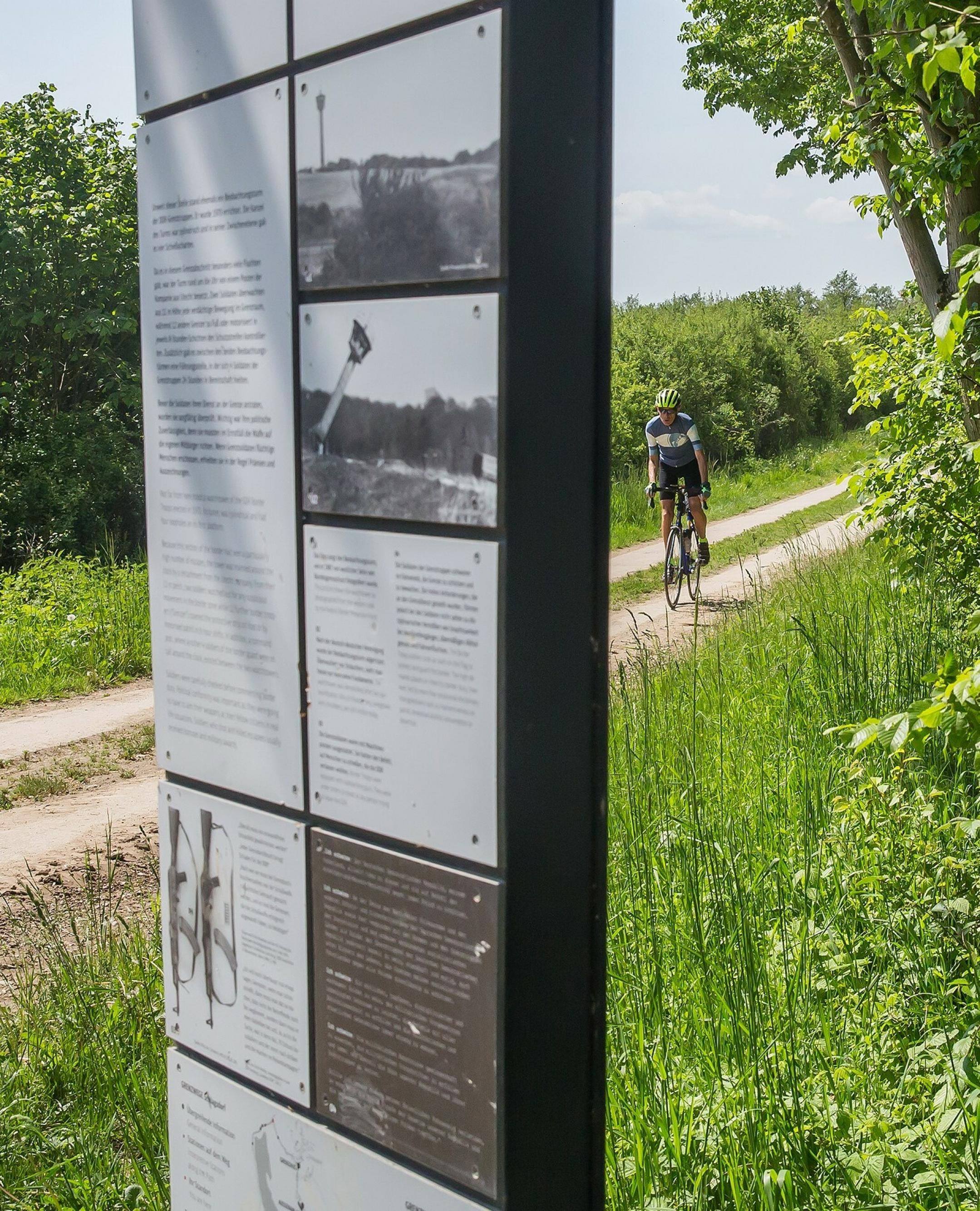 Martin C Roos fährt mit dem Rennrad auf einem alten Kolonnenweg, an dem – im Bildvordergrund – eine Schautafel des Museums zu sehen ist.
