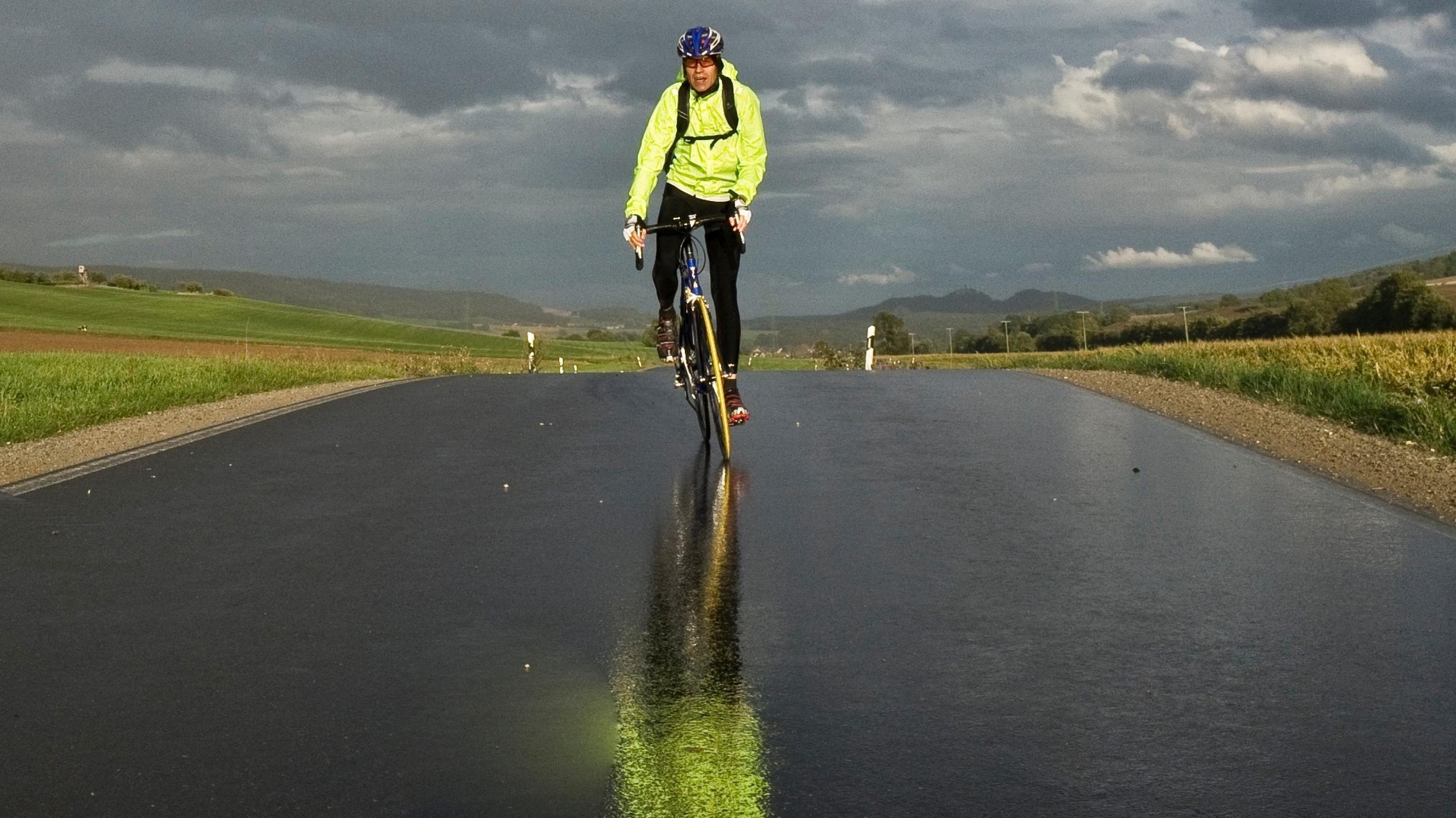 Rennrad-Reporter Martin C Roos fährt auf regennasser Fahrbahn direkt auf die Kamera zu. Die Sonne scheint bereits wieder, am Horizont dräuen dunkle Wolken
