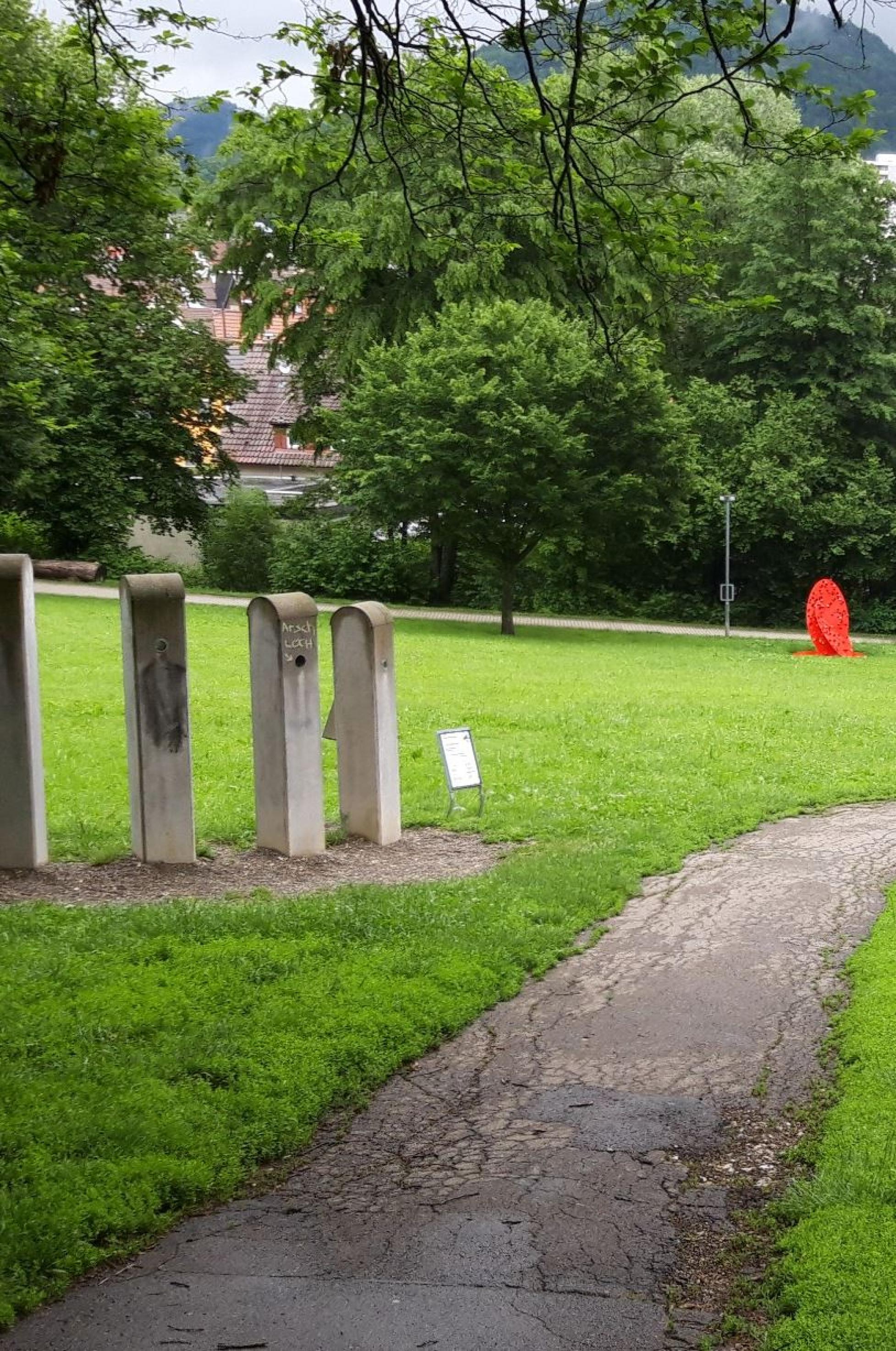 Unverputzte Betonstelen an einem Rasenhang. Der Durchguck einer der Stelen ist beschriftet mi „Arschloch“.