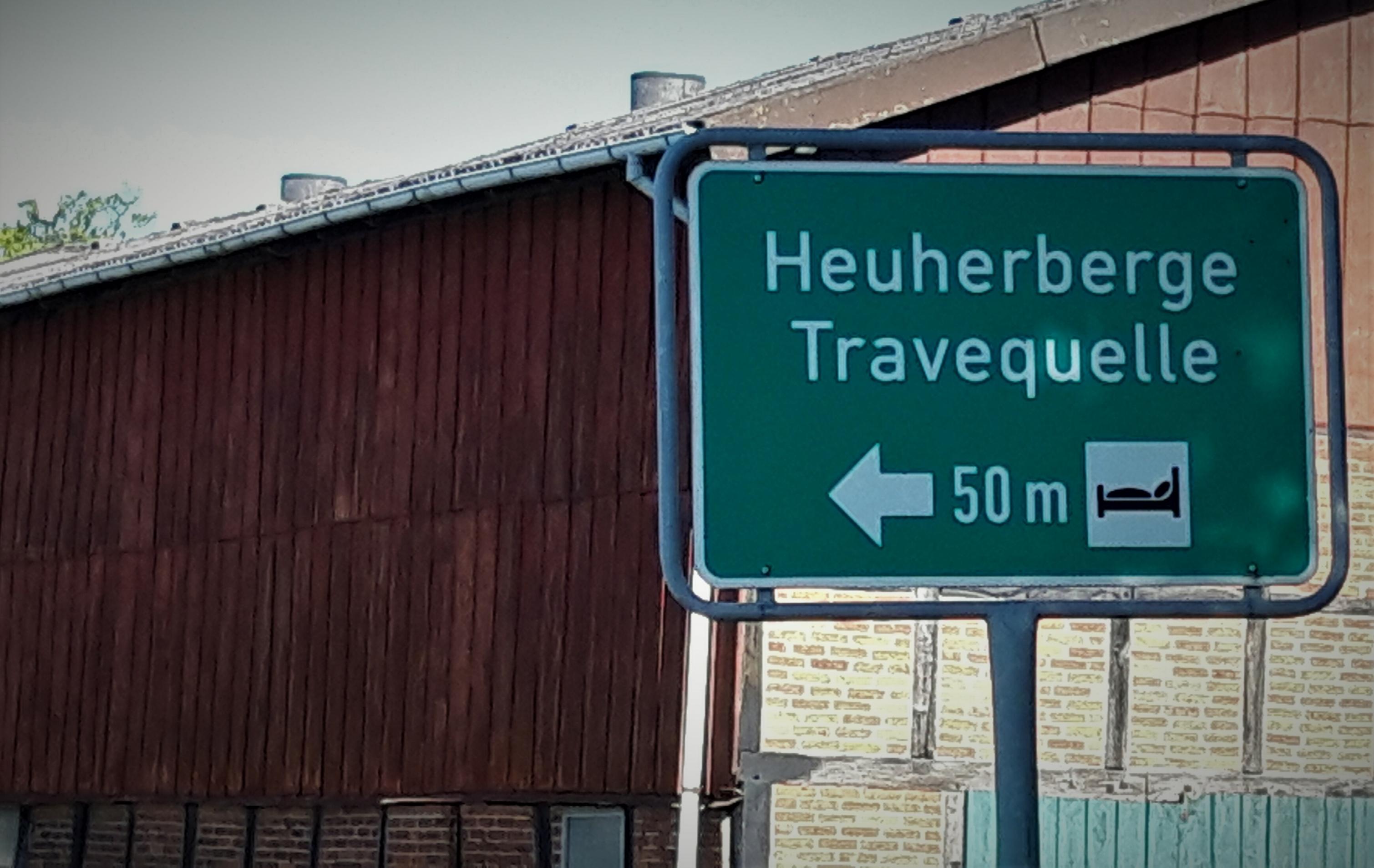 Hinweissschild für die Heuherberge Travequelle, mit Haus im Hintergrund.