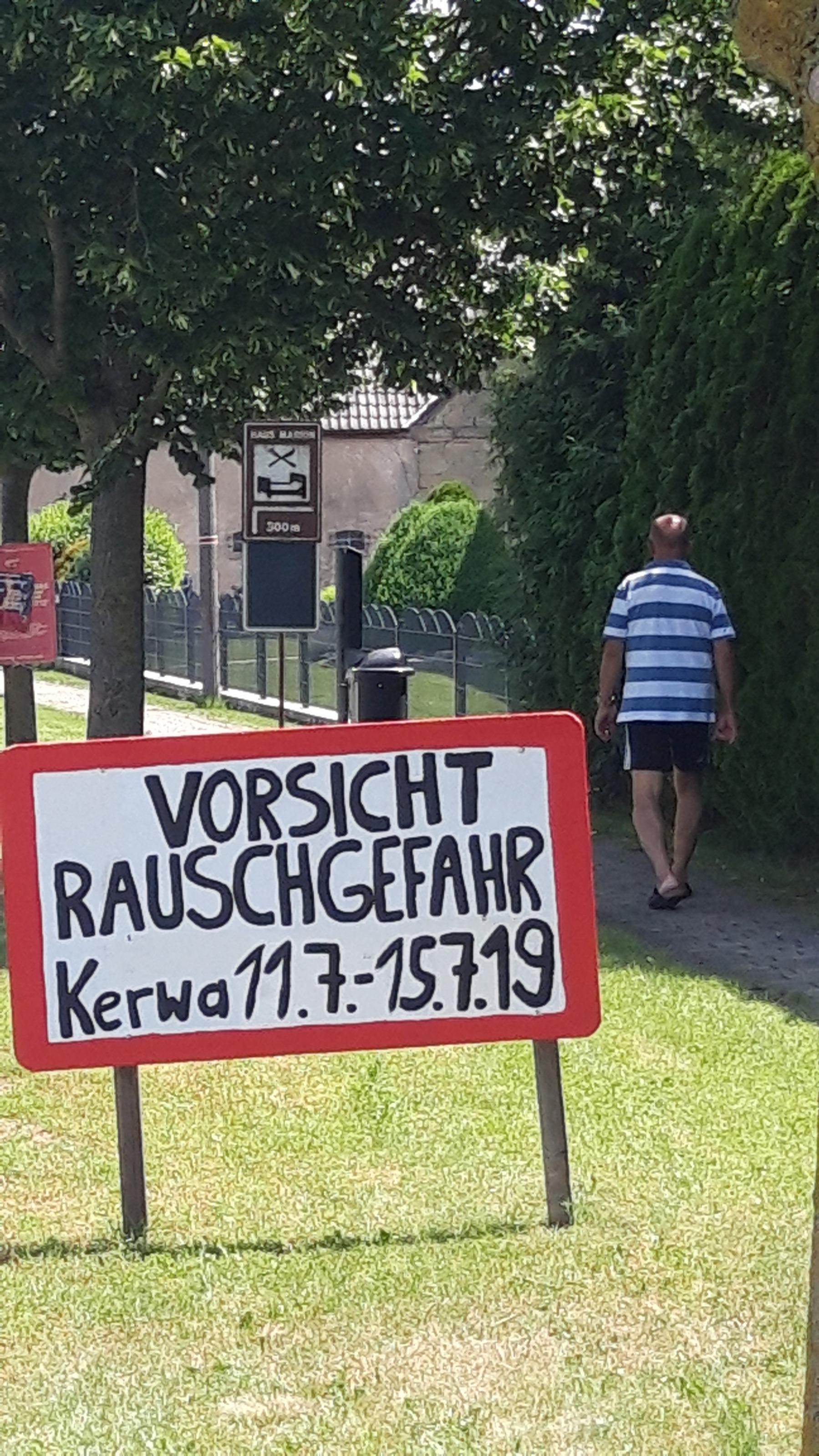 Auf dem Grünstreifen neben der Dorfstraße steht „Vorsicht Rauschgefahr – Kerwa 11.7.-15.7.19“.