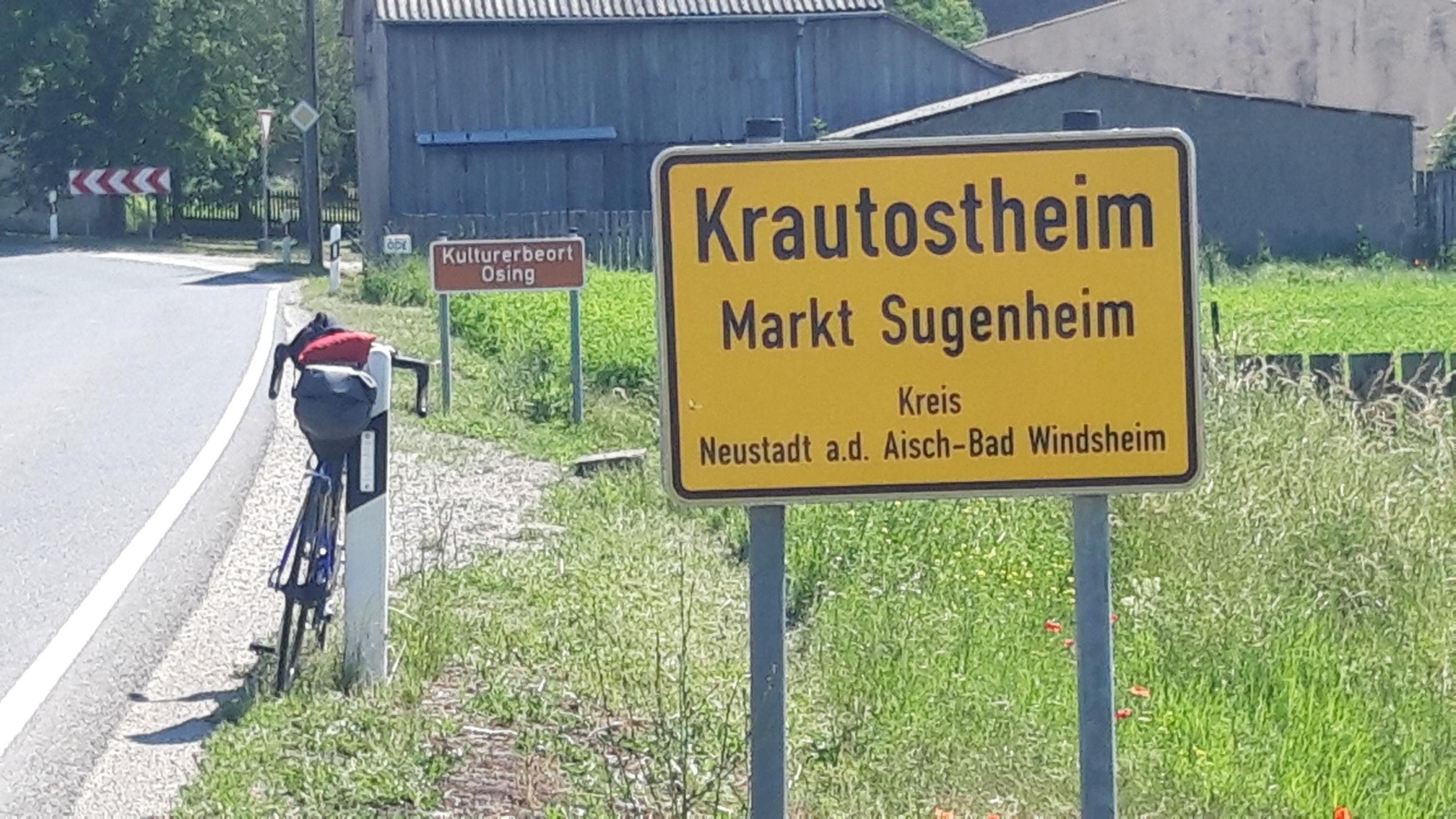 hinter dem Ortschild von Krautostheim ist das braune „Kulturerbe“-Schild zu sehen sowie das Vehikel des RadelndenReporters.