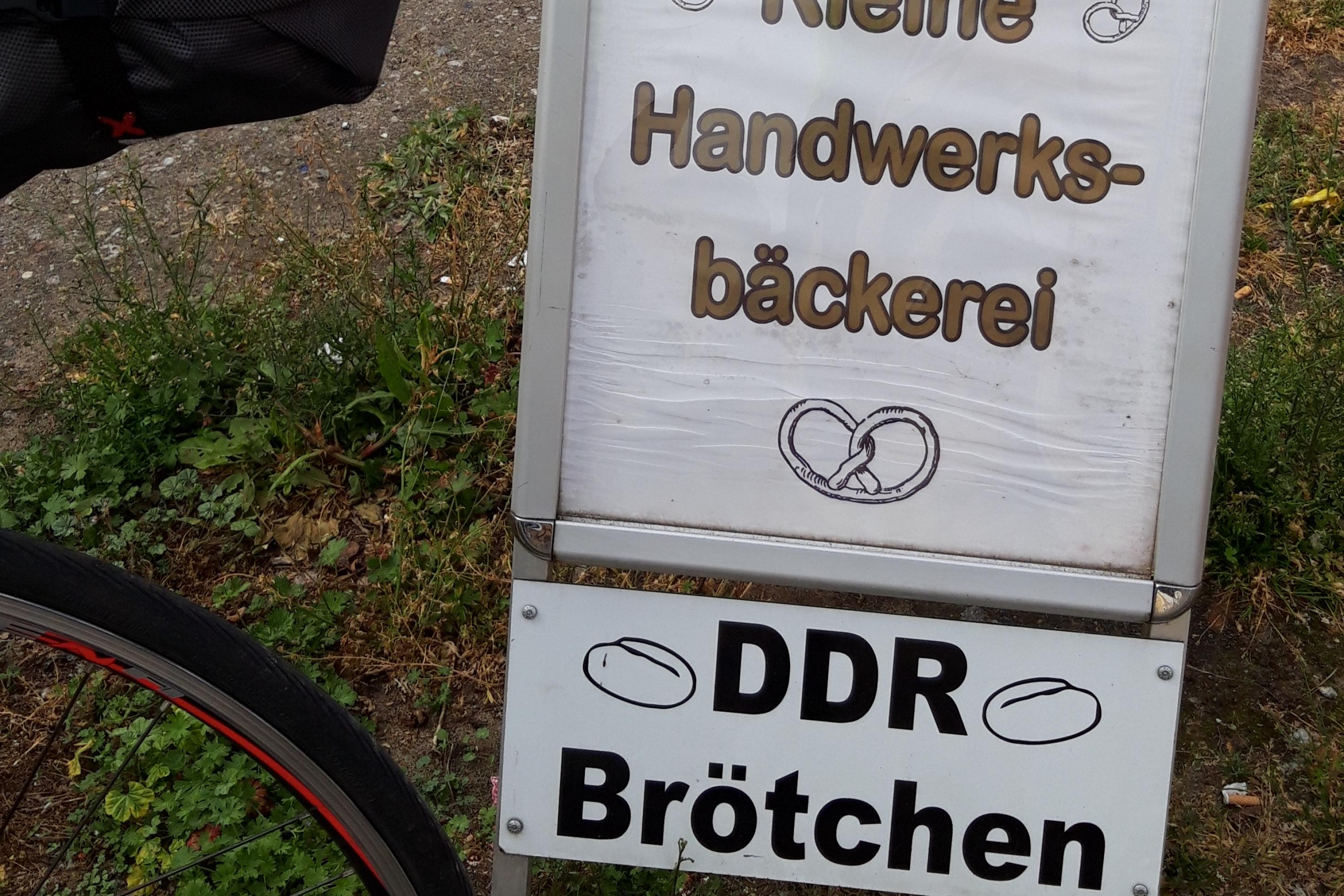 Das Vehikel des RadelndenReporters parkt neben einem Hinweisschild, auf dem zu lesen ist „DDR-Brötchen, Kleine Handwerksbäckerei, 25 Meter“.
